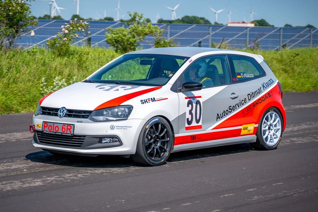 Ein weißer Rennwagen Volkswagen Polo 6R mit roten, schwarzen und blauen Akzenten, Nummer 30, auf einer Rennstrecke, mit Windkraftanlagen im Hintergrund.