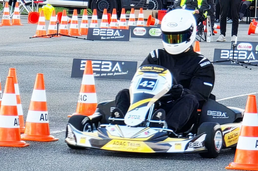 Ein Rennfahrer in Helm und schwarzer Schutzkleidung fährt auf einem Mini-Kart zwischen orangefarbenen Verkehrskegeln bei einem Motorsport-Event.