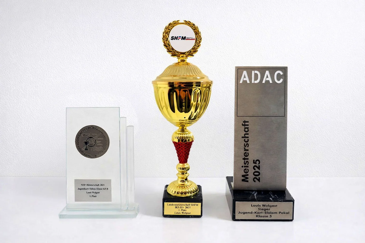 Preise, darunter eine Trophäe, eine transparente Trophäe und eine metallene Trophäe, die alle für Wettbewerbe im Jugendarbeit im Kart-Slalom 2025, Klasse 5, an den Fahrer Louis Wolgast vergeben wurden.