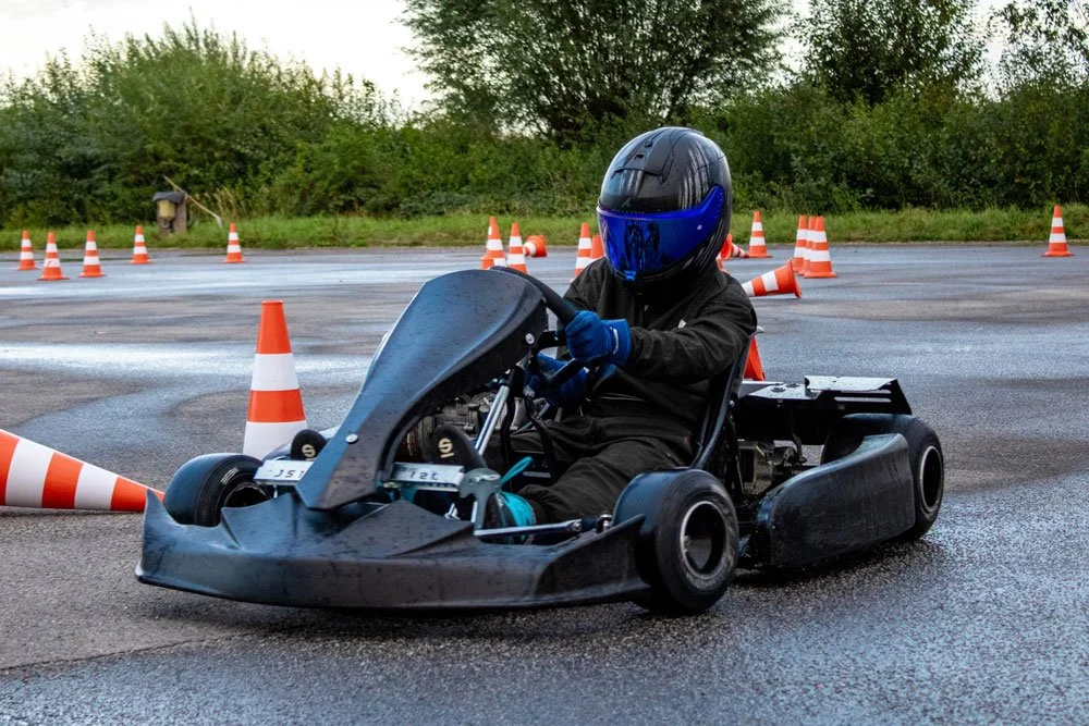 Ein Mensch auf einem Go-Kart trägt eine schwarze Schutzkappe mit blauer Visier und trägt einen schwarzen Anzug, während er auf einer nassen Straße zwischen orangefarbenen Verkehrsabschlusspylonen fährt.