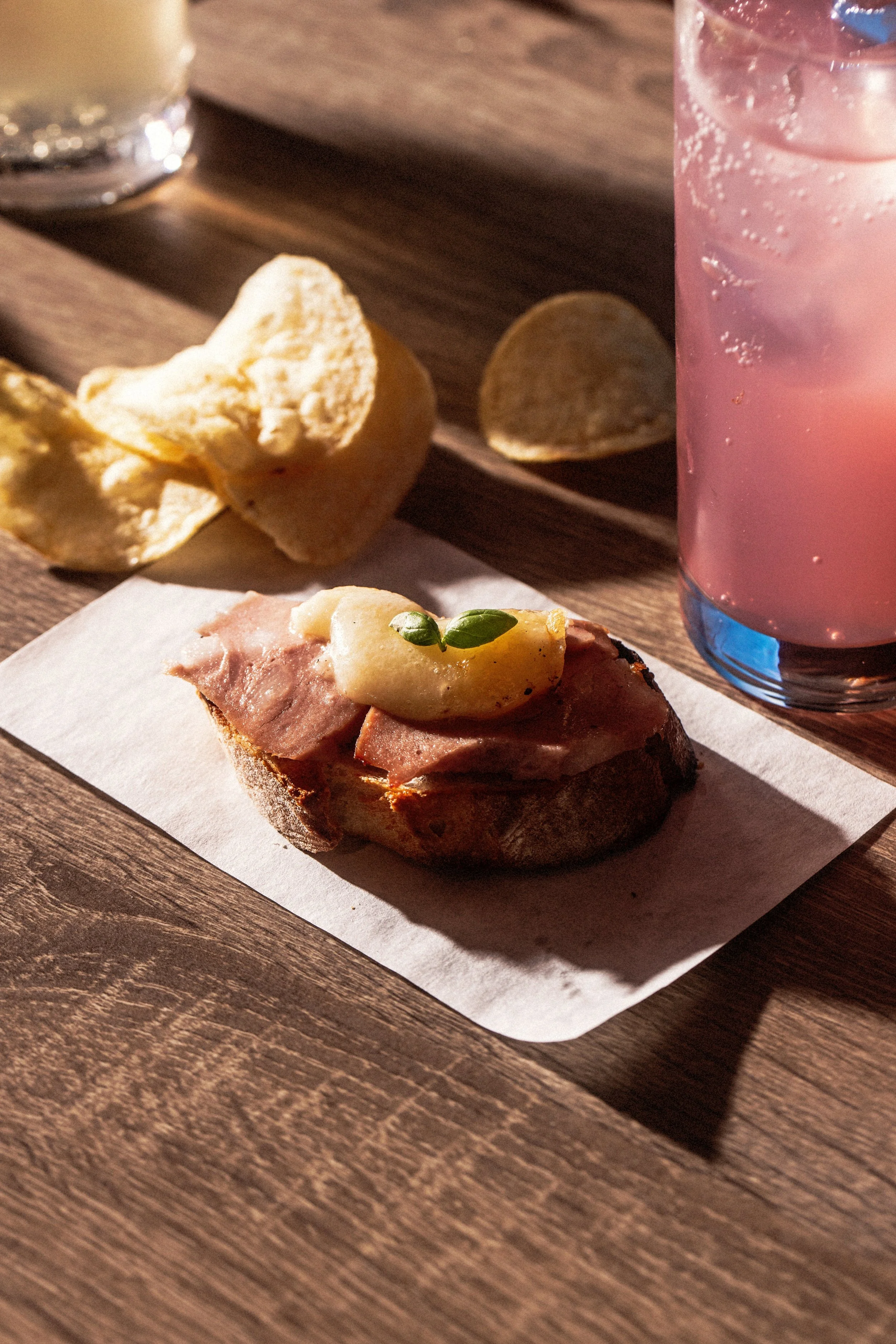 A wooden table with a slice of bread topped with ham and a small sprig of herbs, a piece of bread, a glass of pink soda with ice, and a wooden tray with potato chips.