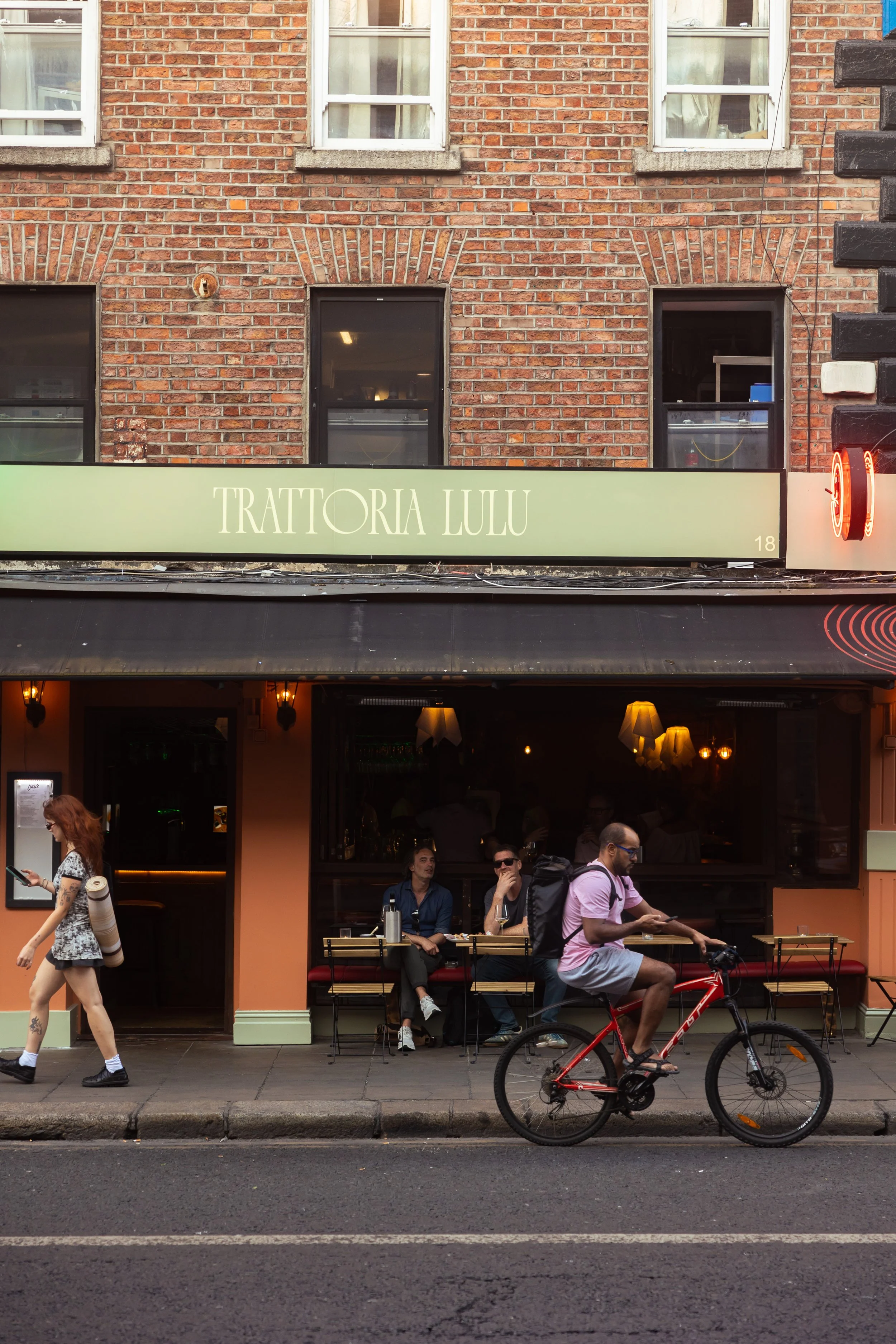 Street view showing a cafe with outdoor seating, people sitting at a table, a man riding a bike, and pedestrians walking by. The cafe has a sign reading 'Trattoria Lulu' on the building's facade.