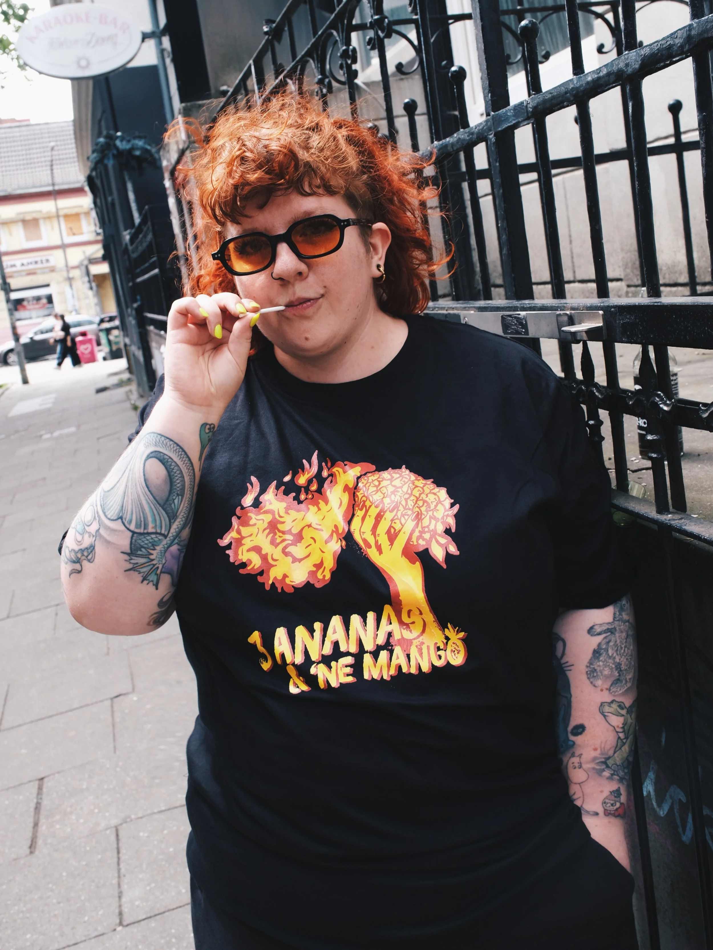 A person with vibrant orange curly hair and tattoos on their arms is sitting outdoors against a black iron fence. They are wearing sunglasses and a black T-shirt with a flaming brain graphic and the text 'Jananas Ne Mango.' They are holding a cigarette near their lips.