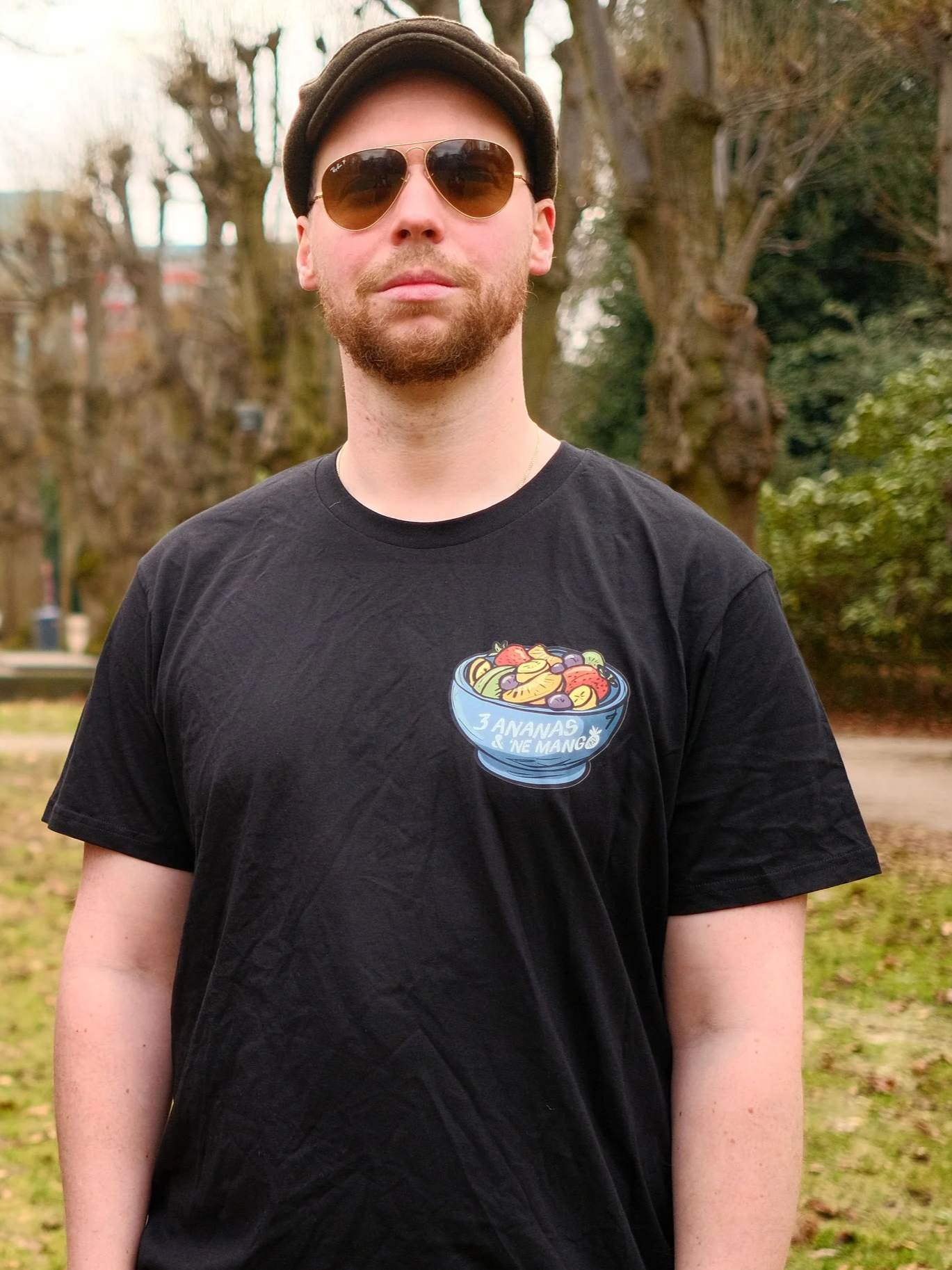 A man standing outdoors in a park with trees in the background. He has a beard, is wearing sunglasses, a brown hat, and a black T-shirt with a colorful fruit bowl graphic and French text that reads '3 Ananas & 1 Mango'.