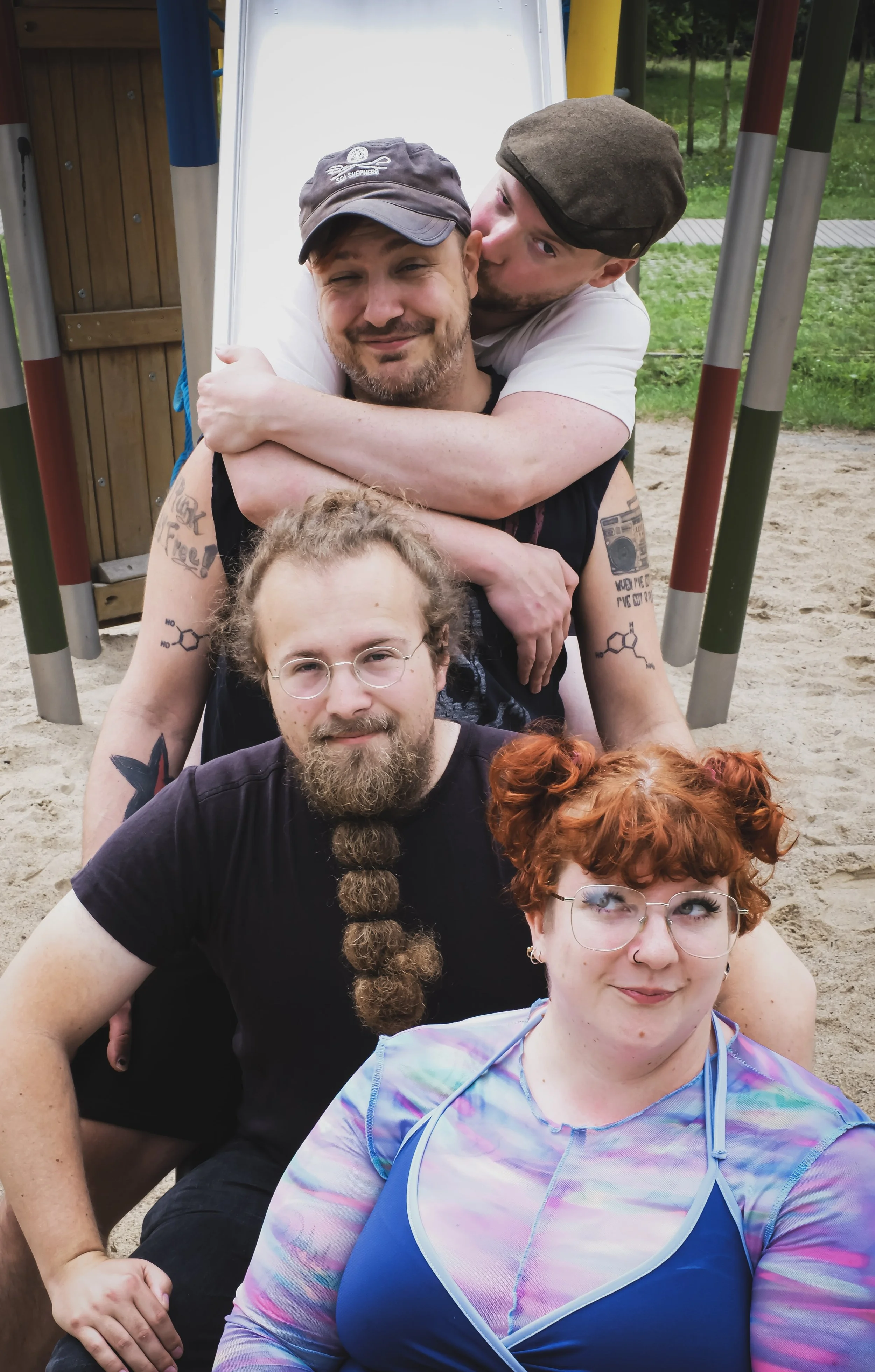 A group of five friends sitting on a playground structure, with three men and two women, smiling and posing for a photo during daytime. One man is hugging another from behind, and the woman in front is looking to her right.