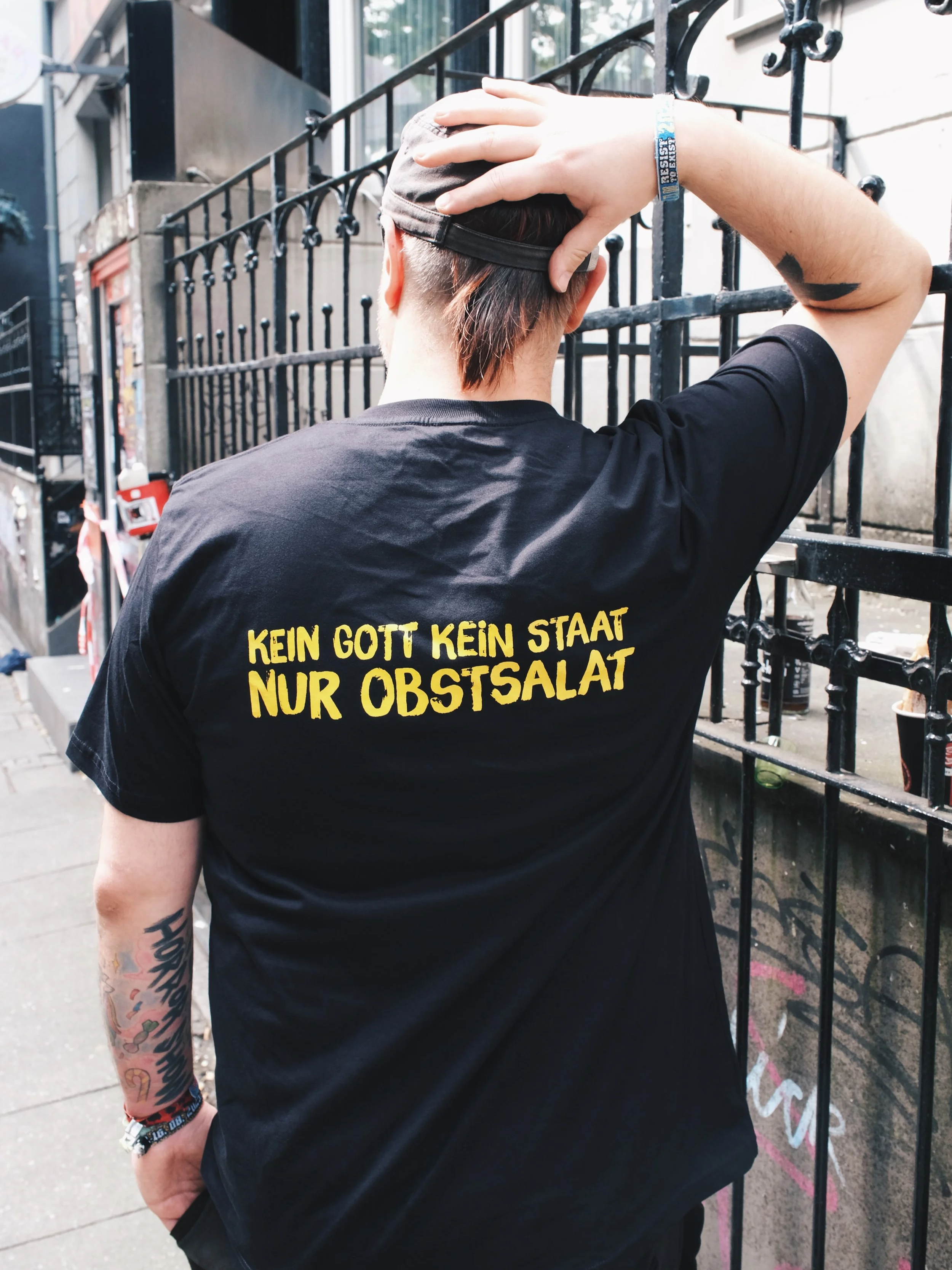 A person standing outdoors in front of a black metal fence, seen from the back, wearing a black t-shirt with yellow text in German and a black cap, touching their head, with colorful tattoos on one arm and a wristband.