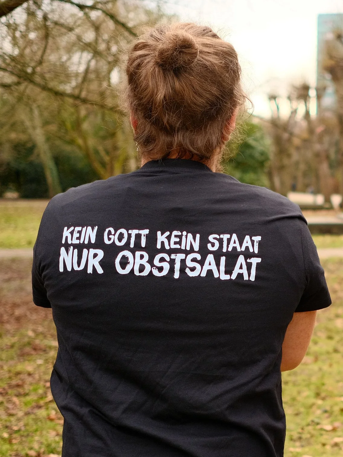 Back view of a woman wearing a black T-shirt with German text, standing outdoors in a park with trees and a blurred background.