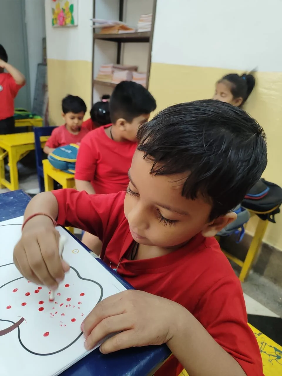 Young boy in a red shirt drawing on a white paper with red dots in a classroom, with other children sitting at yellow desks in the background.
