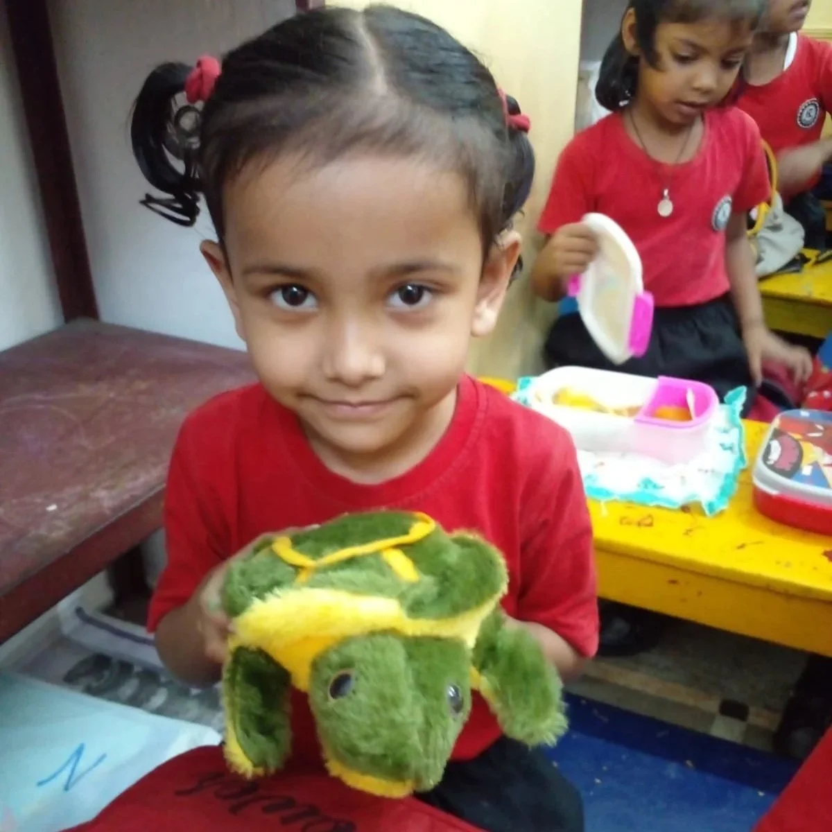 A young girl with dark hair in pigtails holding a plush toy of a colorful chameleon in a classroom setting.