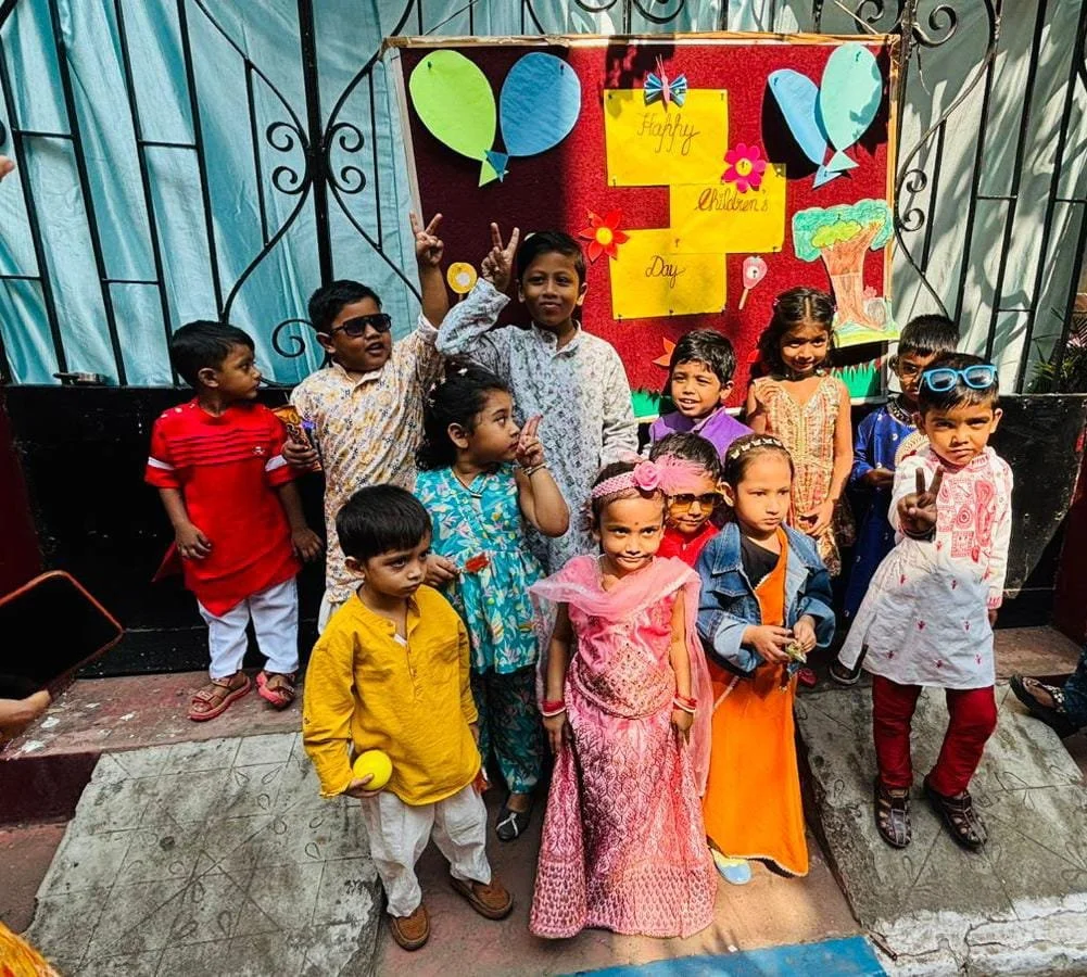 Group of children celebrating Children's Day, dressed in colorful traditional and casual clothing, in front of a decorated red background with balloons and a sign that reads 'Happy Children's Day'.