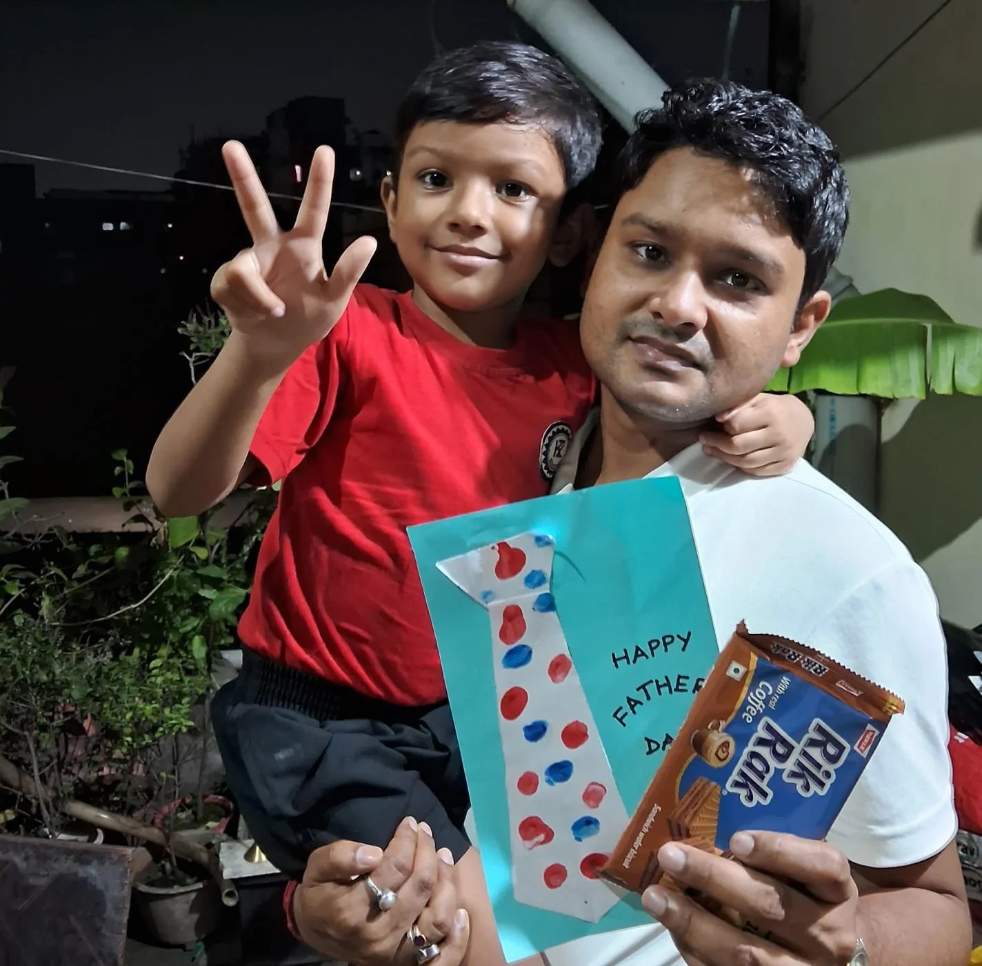 Man and boy celebrating Father's Day, man holding a chocolate snack, boy making a peace sign, boy holding a handmade card that says Happy Father's Day, night setting with plants in background.