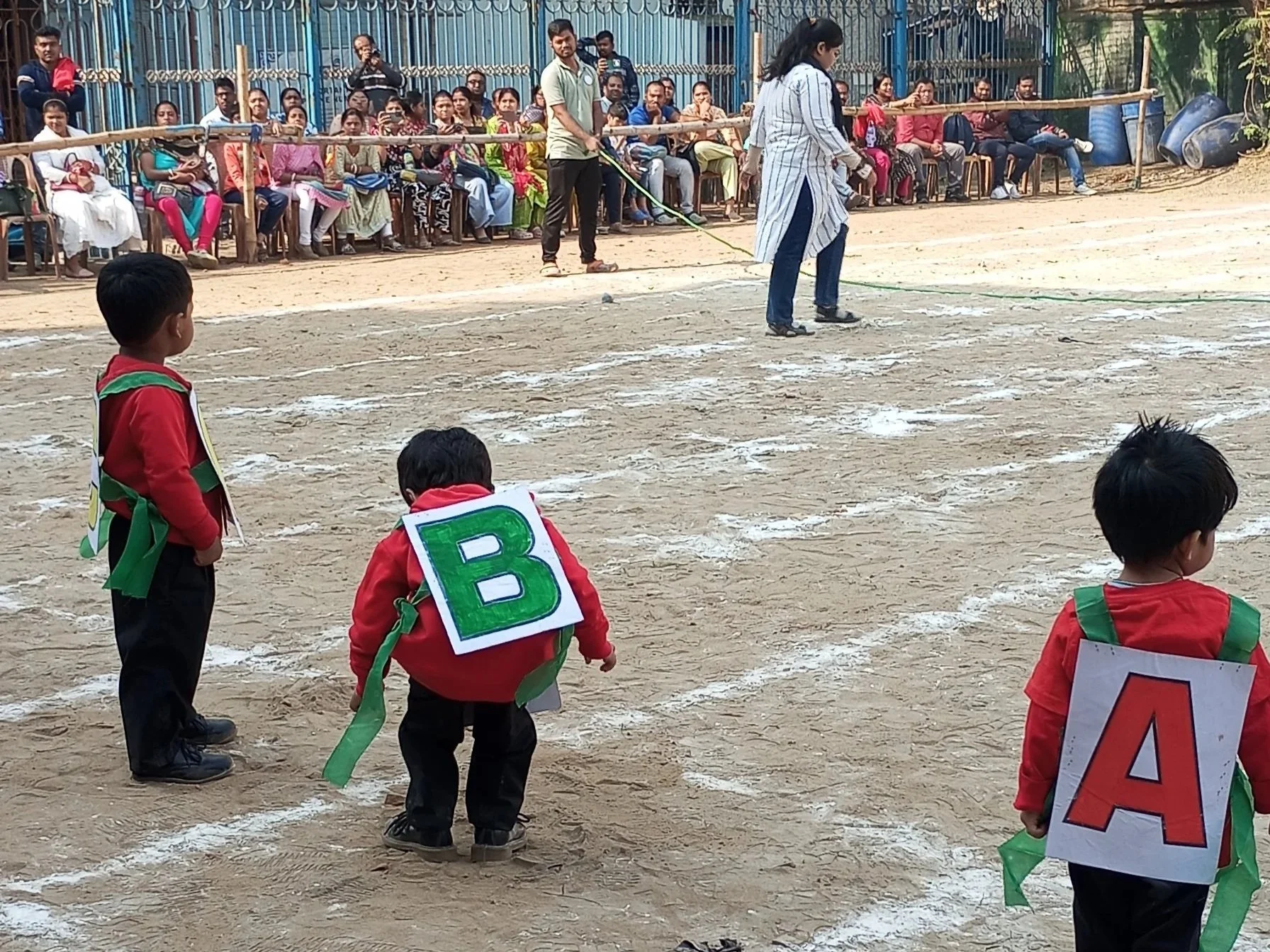 Kids standing on a dirt field with white lines, observing a race, with spectators sitting behind a fence in the background. Some kids are wearing signs with letters, and adults are sitting and standing along the fence.