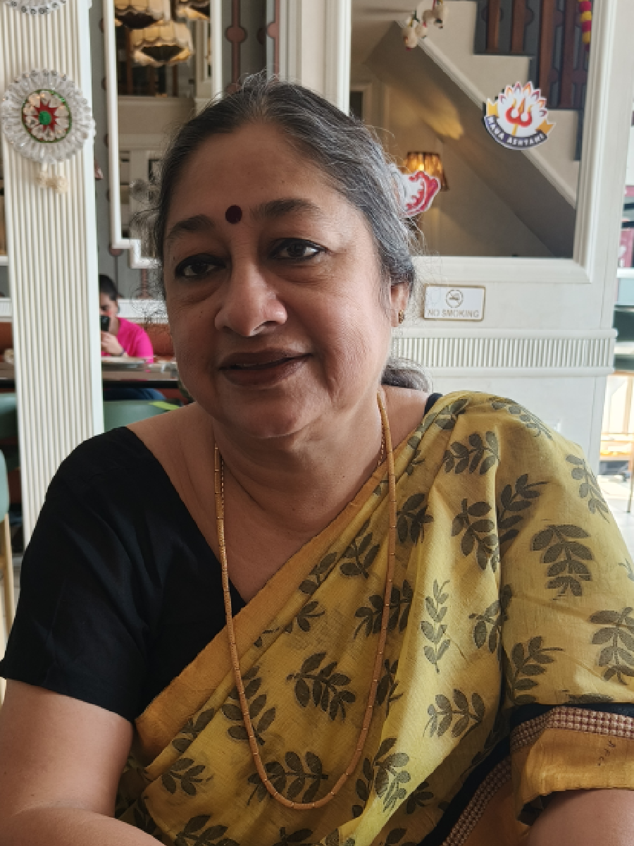 A woman wearing a yellow sari with black leaf patterns, seated indoors, with decorations and a staircase in the background.