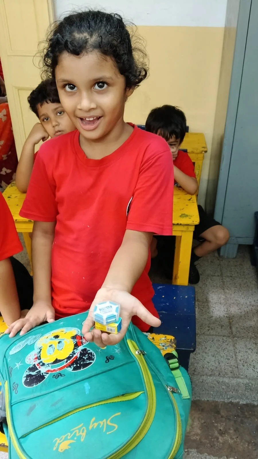 A smiling young girl with curly hair in a red shirt is holding a small item and standing in front of a school bag with a Mickey Mouse design. Behind her are three boys, two sitting at yellow desks and one peeking from behind the girl.