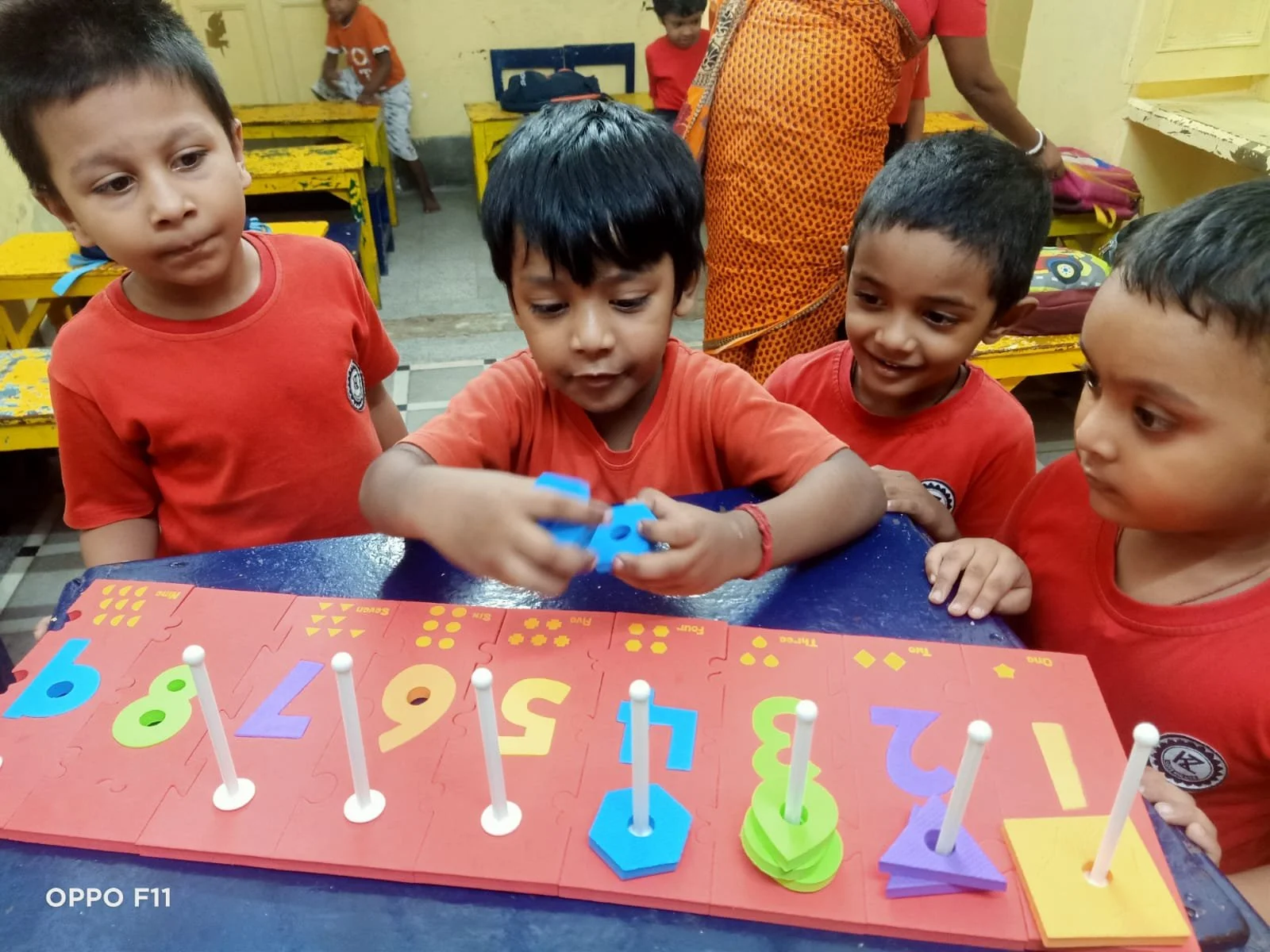 Four young boys in red shirts are gathered around a table, playing with a colorful educational toy that has numbered puzzle pieces and vertical pegs. One boy in the center is placing a piece on the table, while the others watch with interest in a classroom setting.