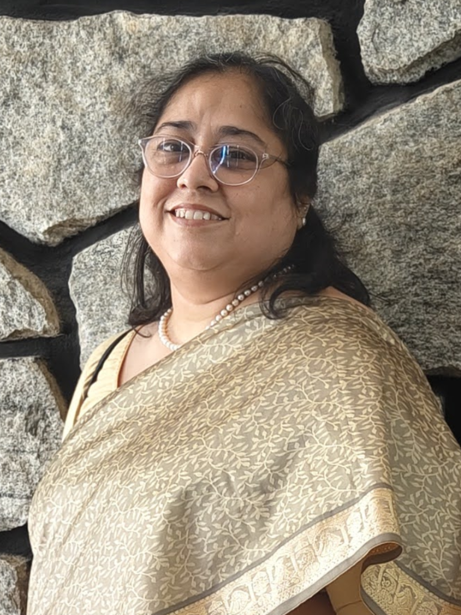 A smiling woman with glasses, wearing a pearl necklace and a patterned saree, standing against a stone wall.