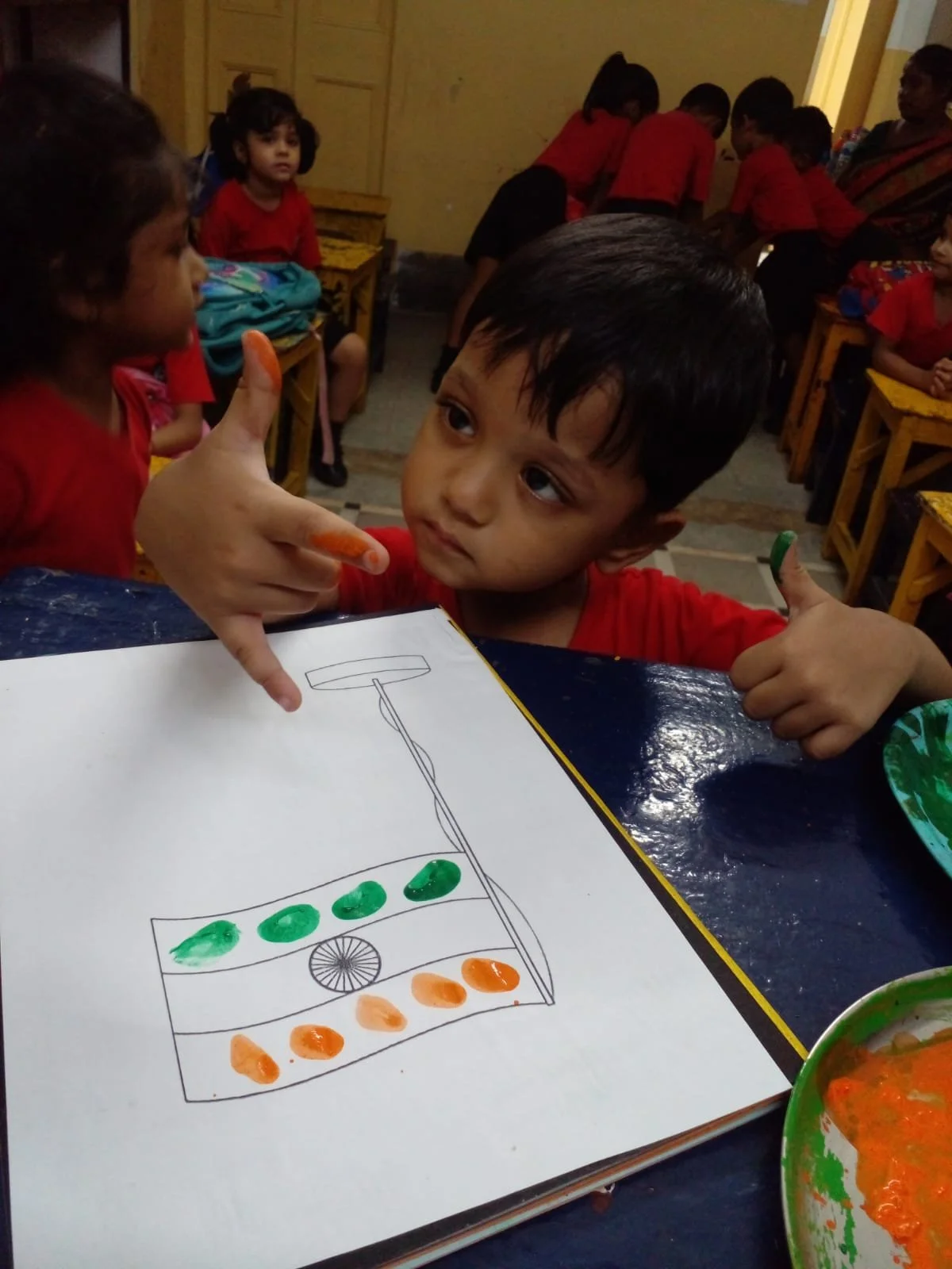 Young boy in a classroom coloring a drawing of a traffic light on a white sheet of paper. He's showing a thumbs-up and has colorful spots of paint on his fingers. Other children in red outfits sit and work at desks in the background.