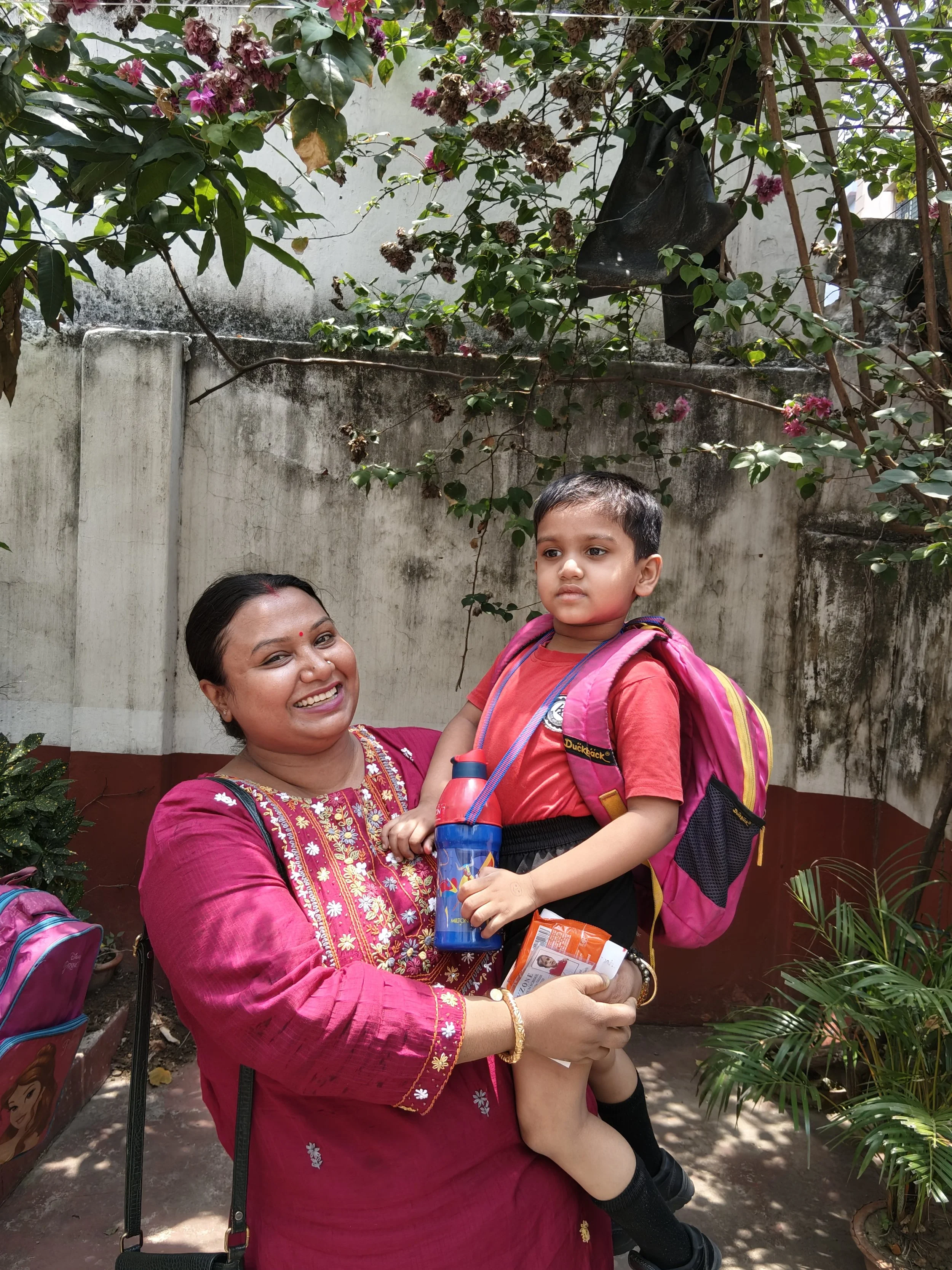 A woman wearing a red traditional outfit is holding a young boy in a pink backpack and red shirt. The woman is smiling at the camera, and the boy looks slightly away. They are standing outdoors in front of a white wall with flowering plants and greenery around.