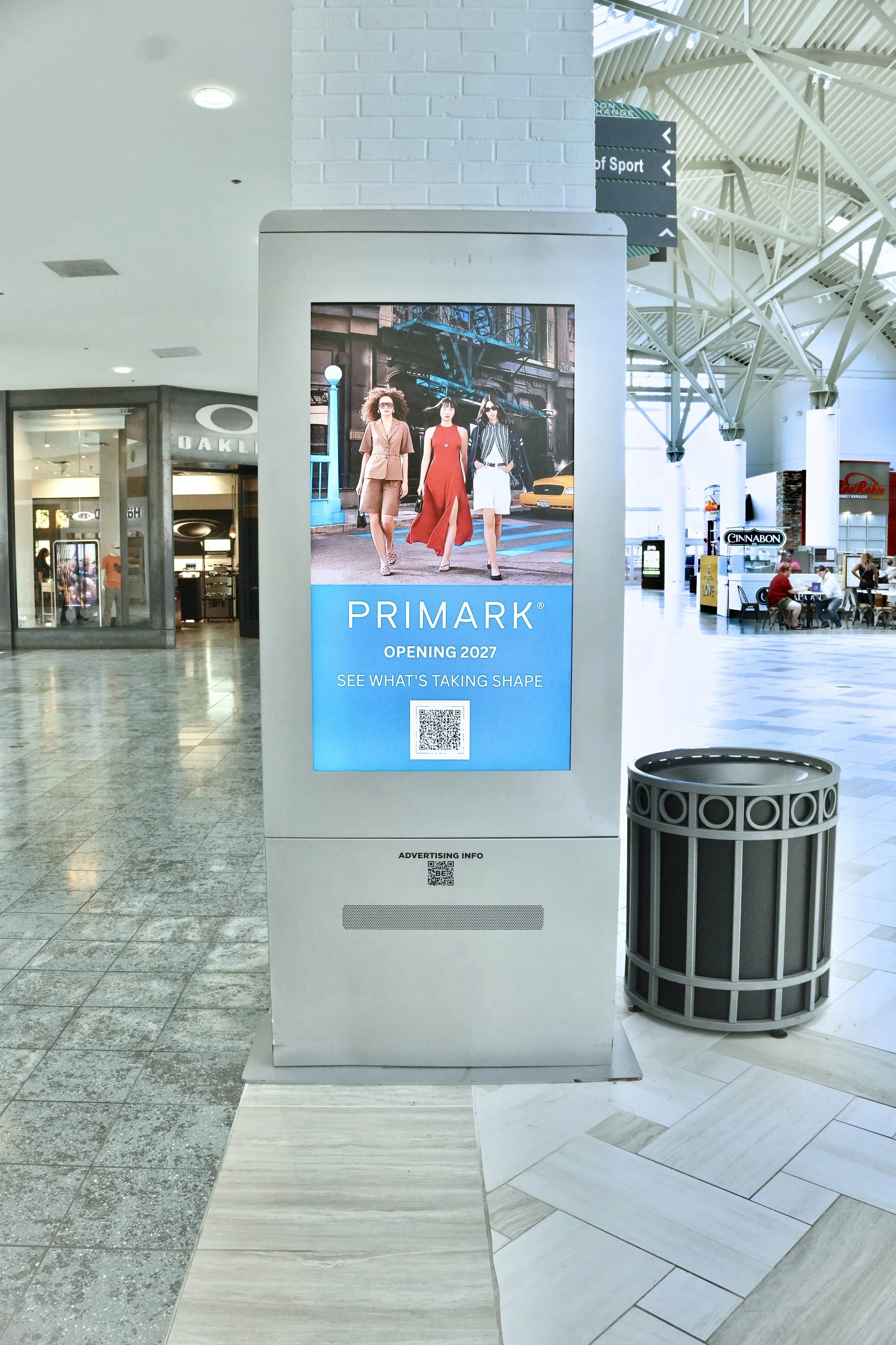 Digital advertisement board showing a picture of three fashionable women walking on a city street, with text below reading "PRIMARK OPENING 2027 SEE WHAT'S TAKING SHAPE" and a QR code underneath, located inside a shopping mall.