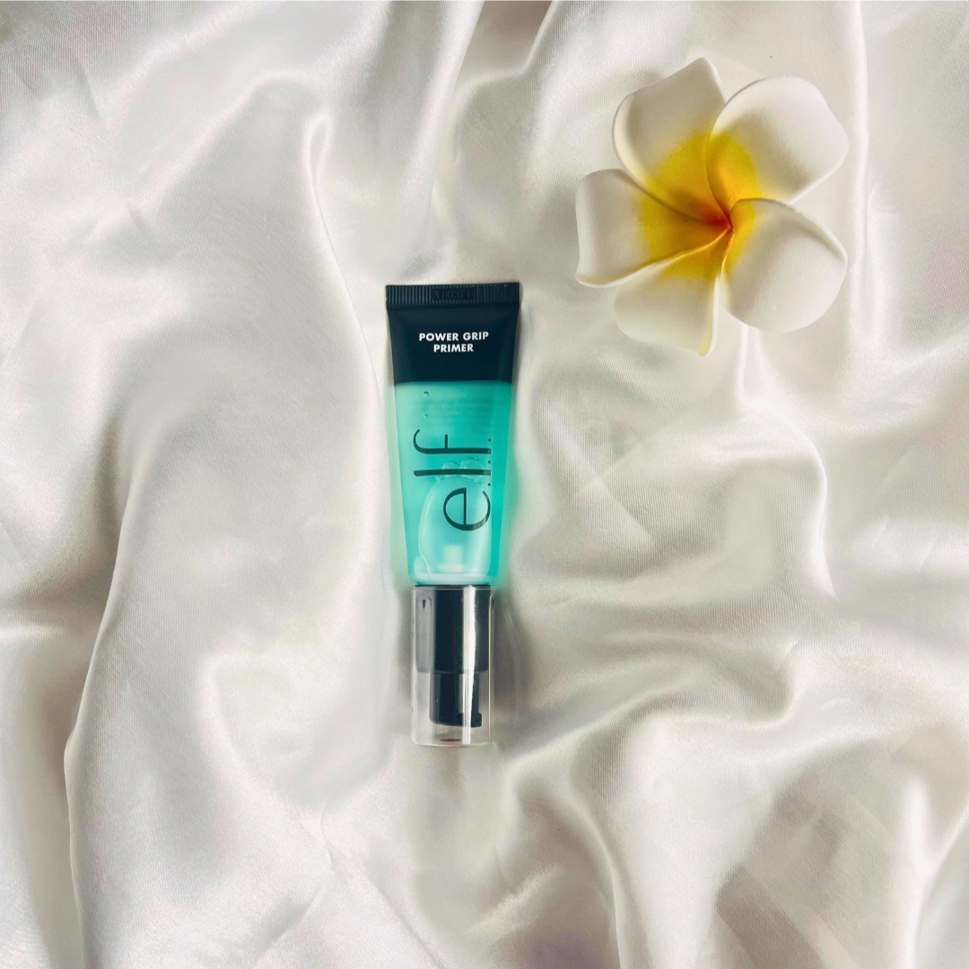 A small tube of e.l.f. Power Grip Primer displaying a gradient of turquoise to clear on a white satin fabric, accompanied by a white and yellow plumeria flower.