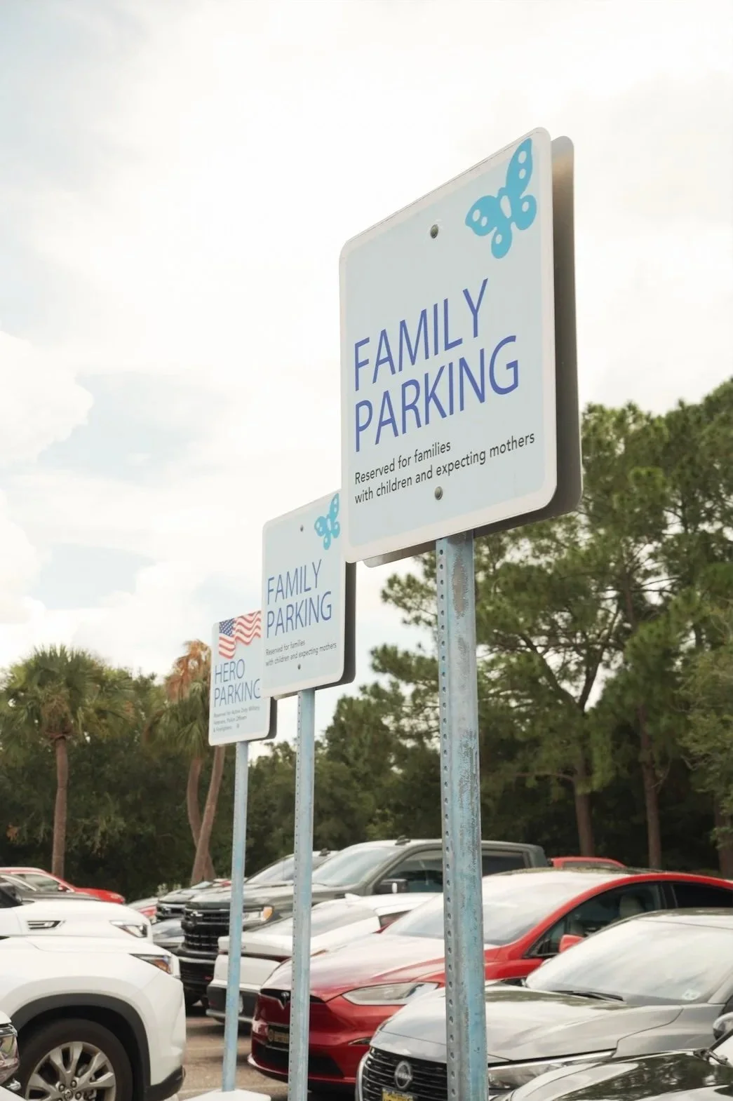 Parking lot with signs for family, hero, and other parking, surrounded by trees and parked cars.