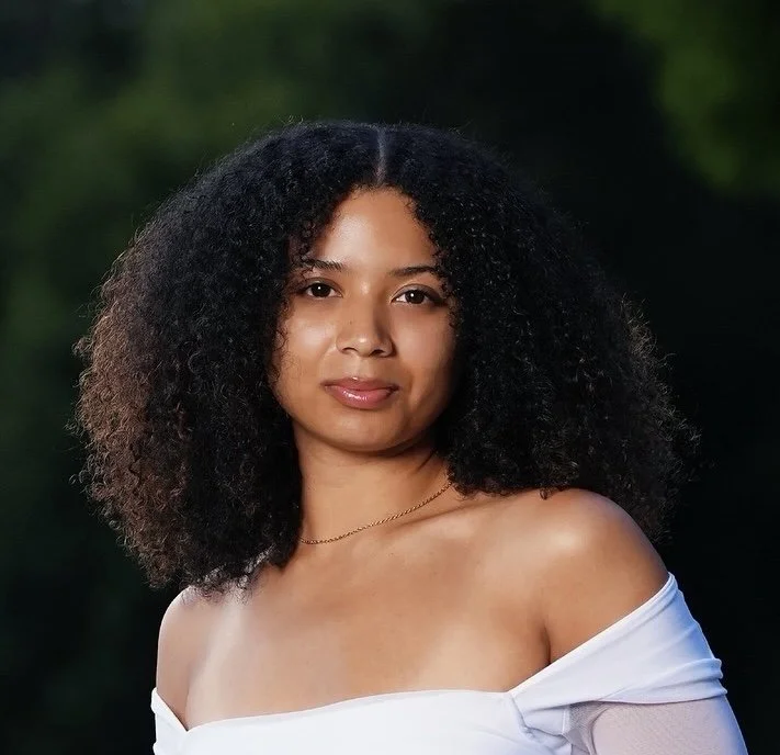 A woman with curly black hair and wearing a white off-the-shoulder top, standing outdoors with a blurred green background.