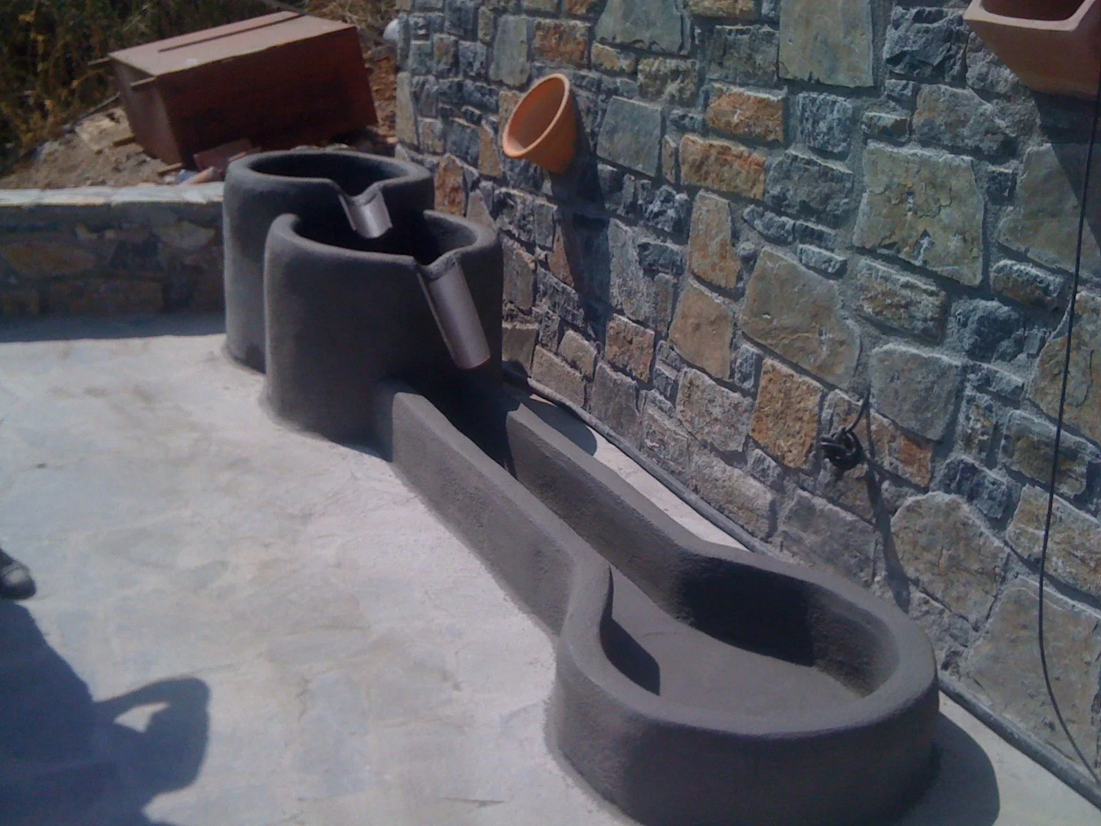 An outdoor scene featuring a concrete skateboard ramp, a stone wall with a terracotta flowerpot, a black and gray skateboarding helmet, and a black wired extension cord attached to the wall.