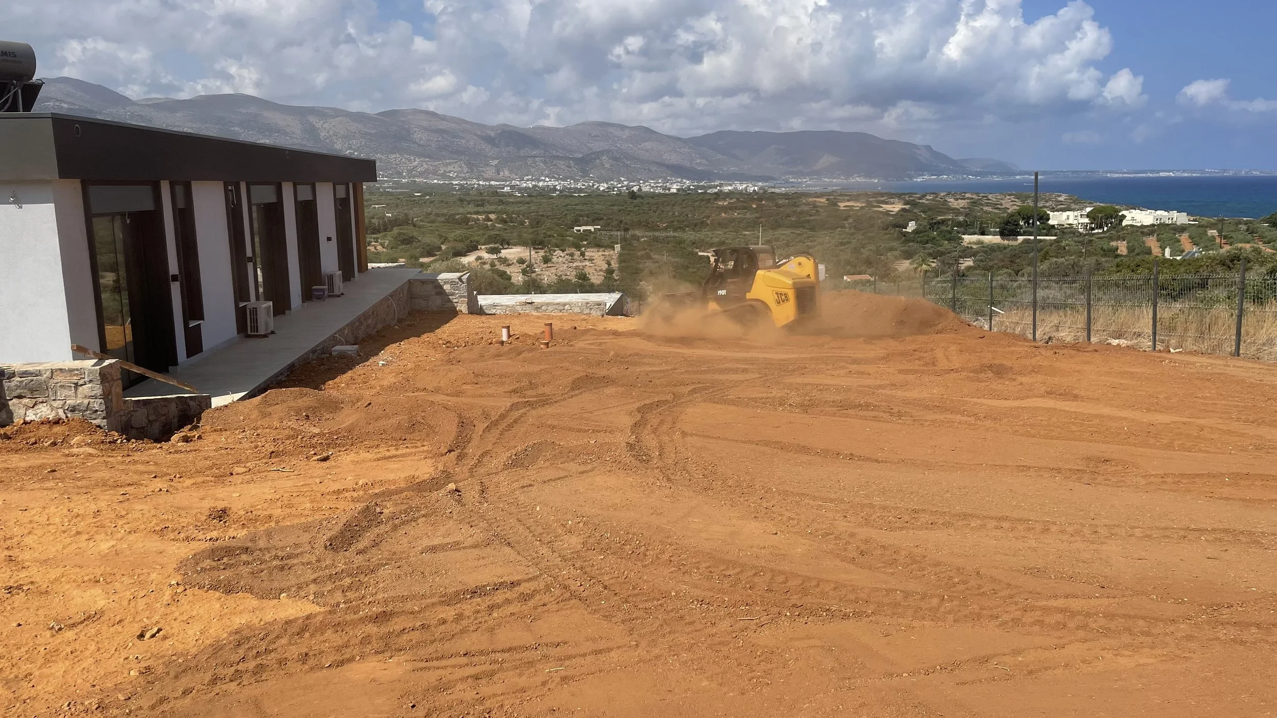 A construction site with a yellow JCB mini excavator working on red dirt, overlooking a landscape with houses, greenery, and mountains in the background, near the coastline.