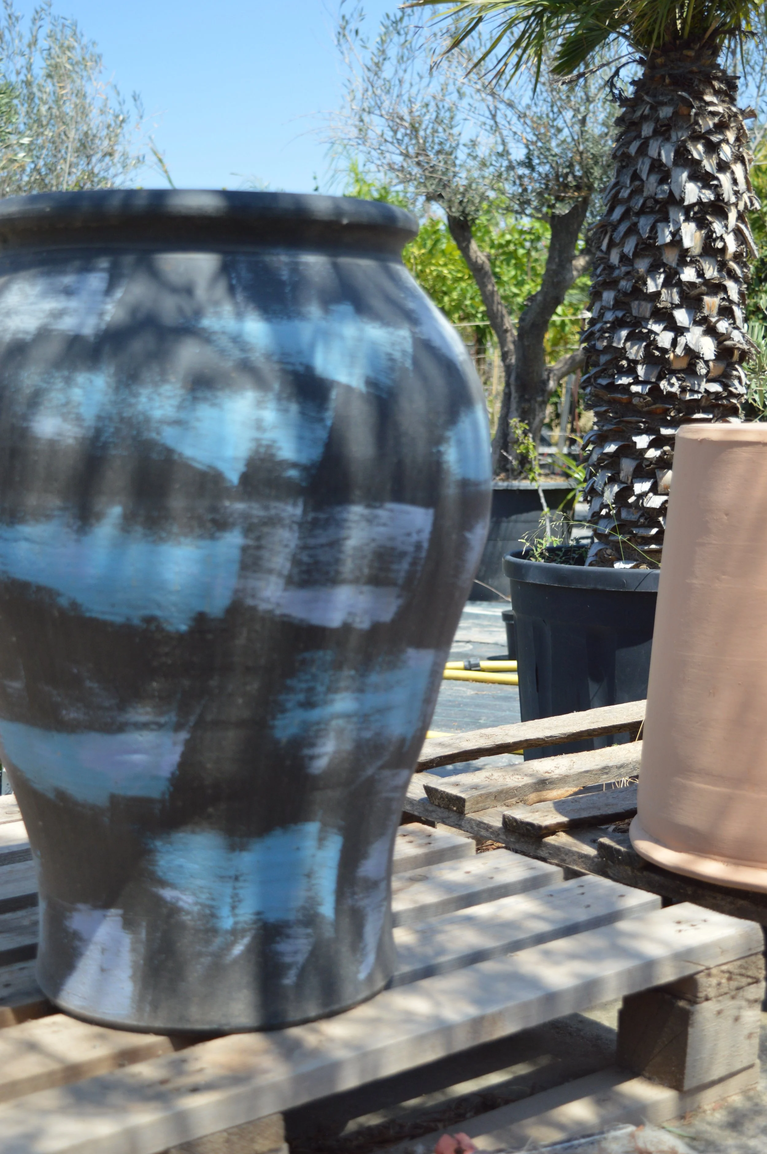 Close-up of a black and white painted ceramic pot on a wooden pallet, with fruit trees in the background under a clear blue sky.
