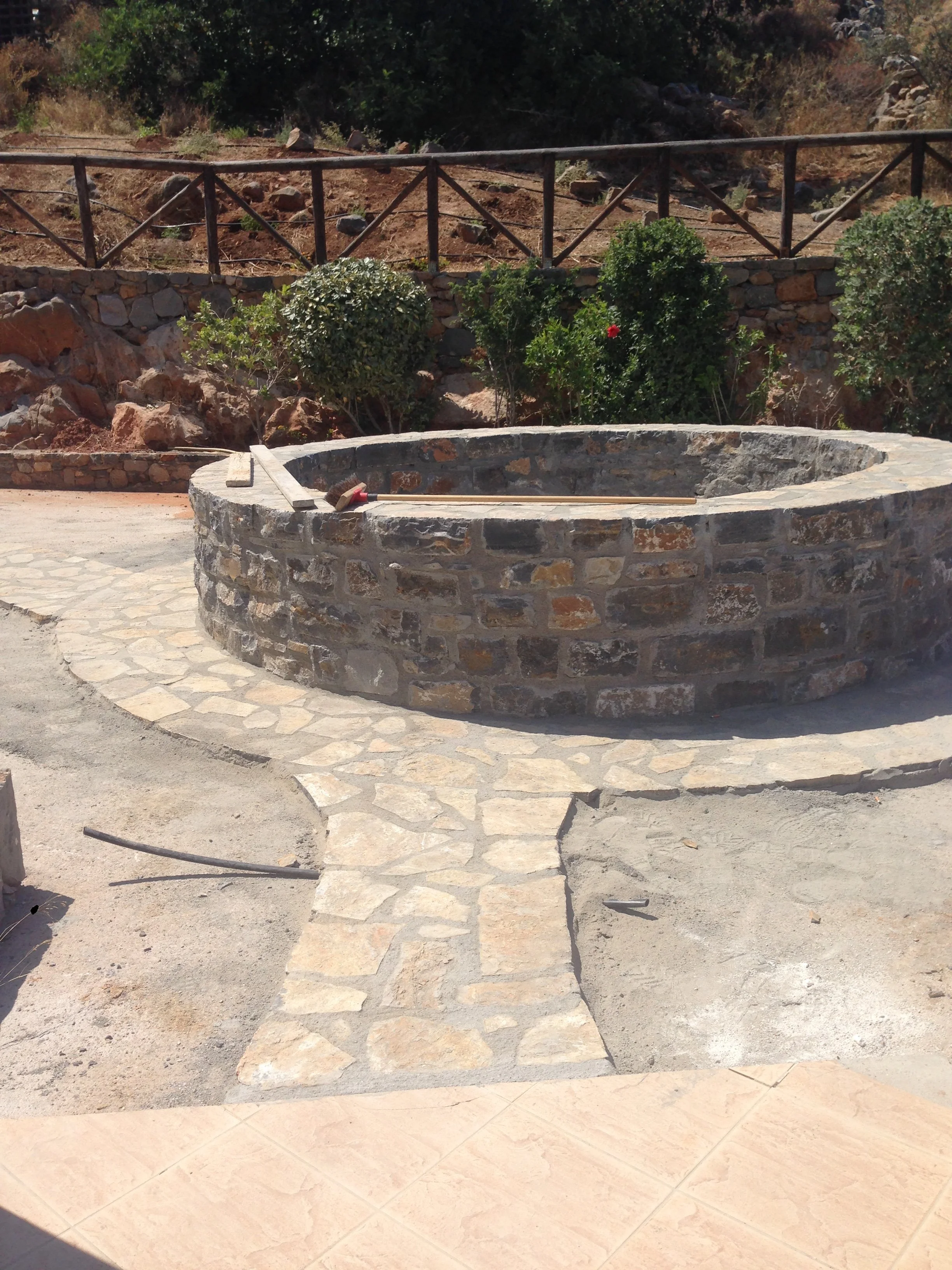 A circular stone fire pit with a long wooden stick and a hammer resting on top, surrounded by stone paving, with a backdrop of bushes, rocks, and a hillside with dry dirt and sparse vegetation.