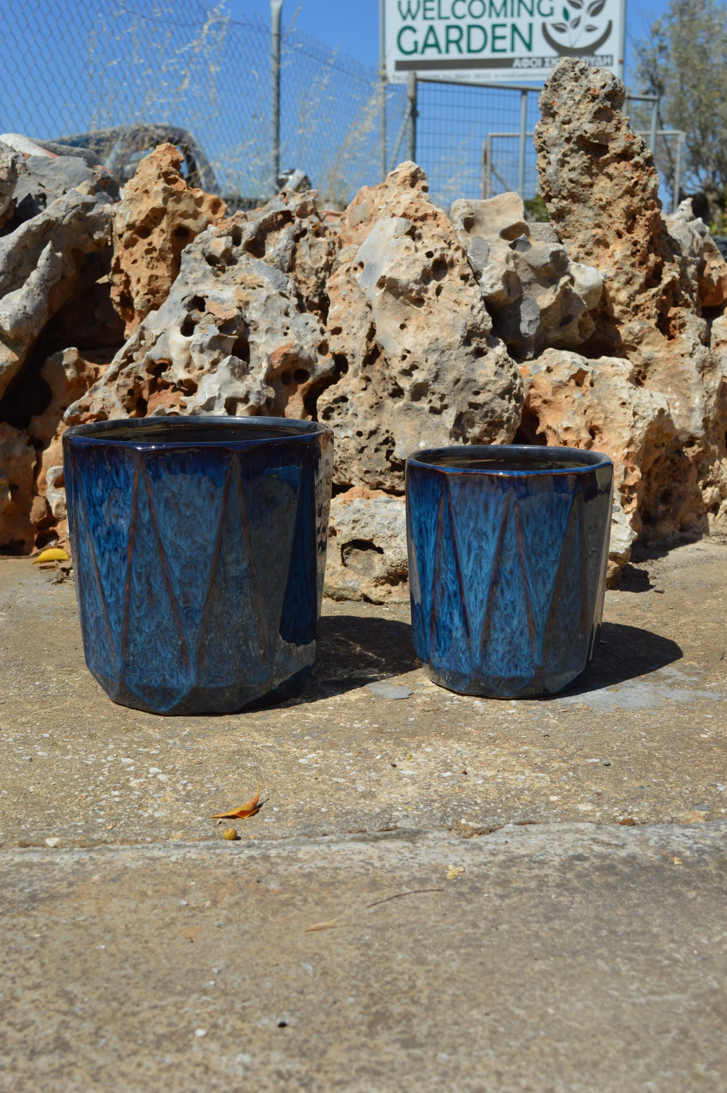 Two dark blue ceramic cups with a marbled pattern sit on a rough concrete surface. Behind them, a pile of large, irregularly shaped rocks or concrete debris is visible. In the background, there is a chain-link fence with a sign that reads 'Welcoming 