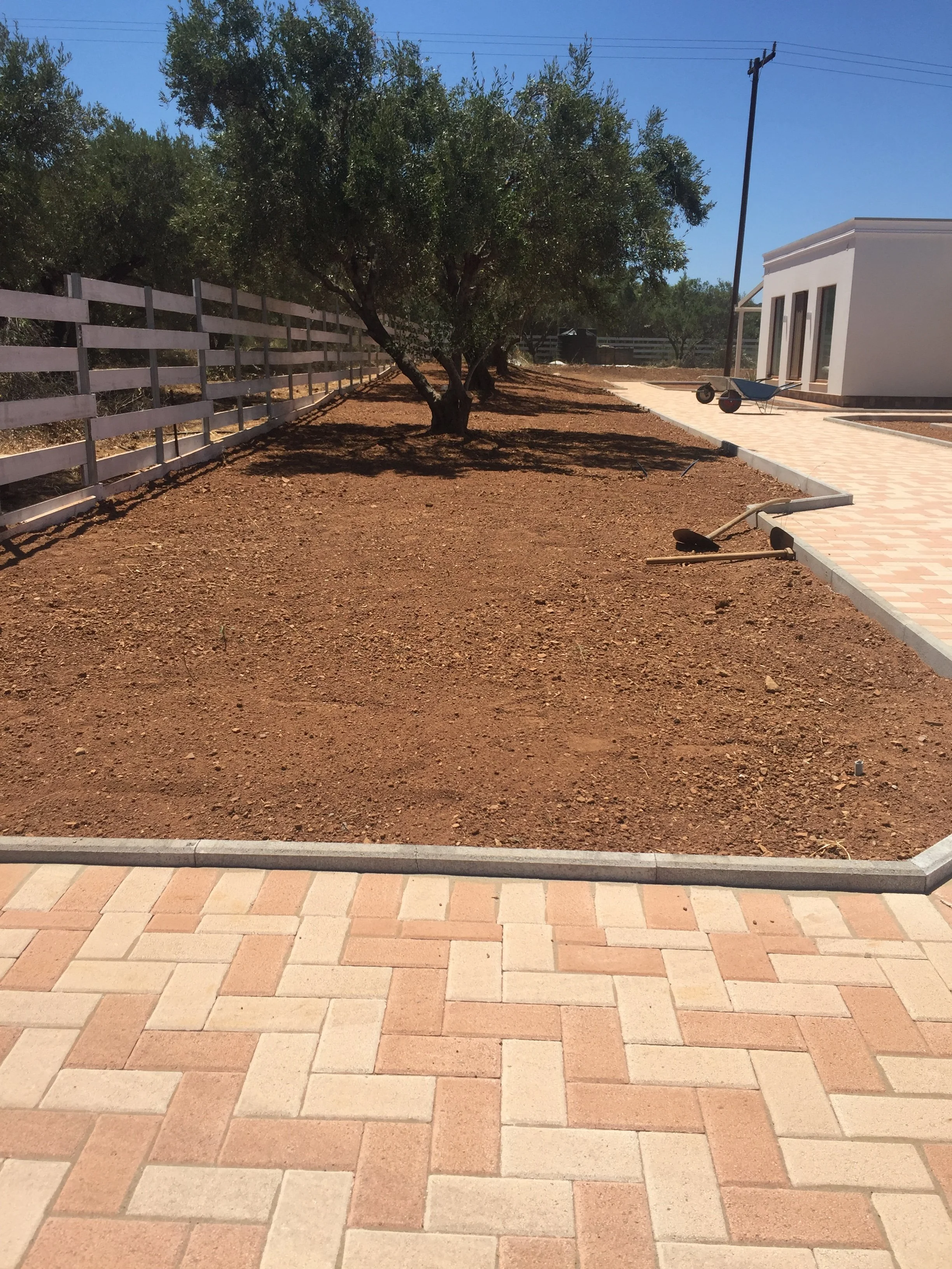 Construction or landscaping site with a partially paved patio area, bare soil, a white building, a wheelbarrow, and a spade. Trees and a fence are in the background.