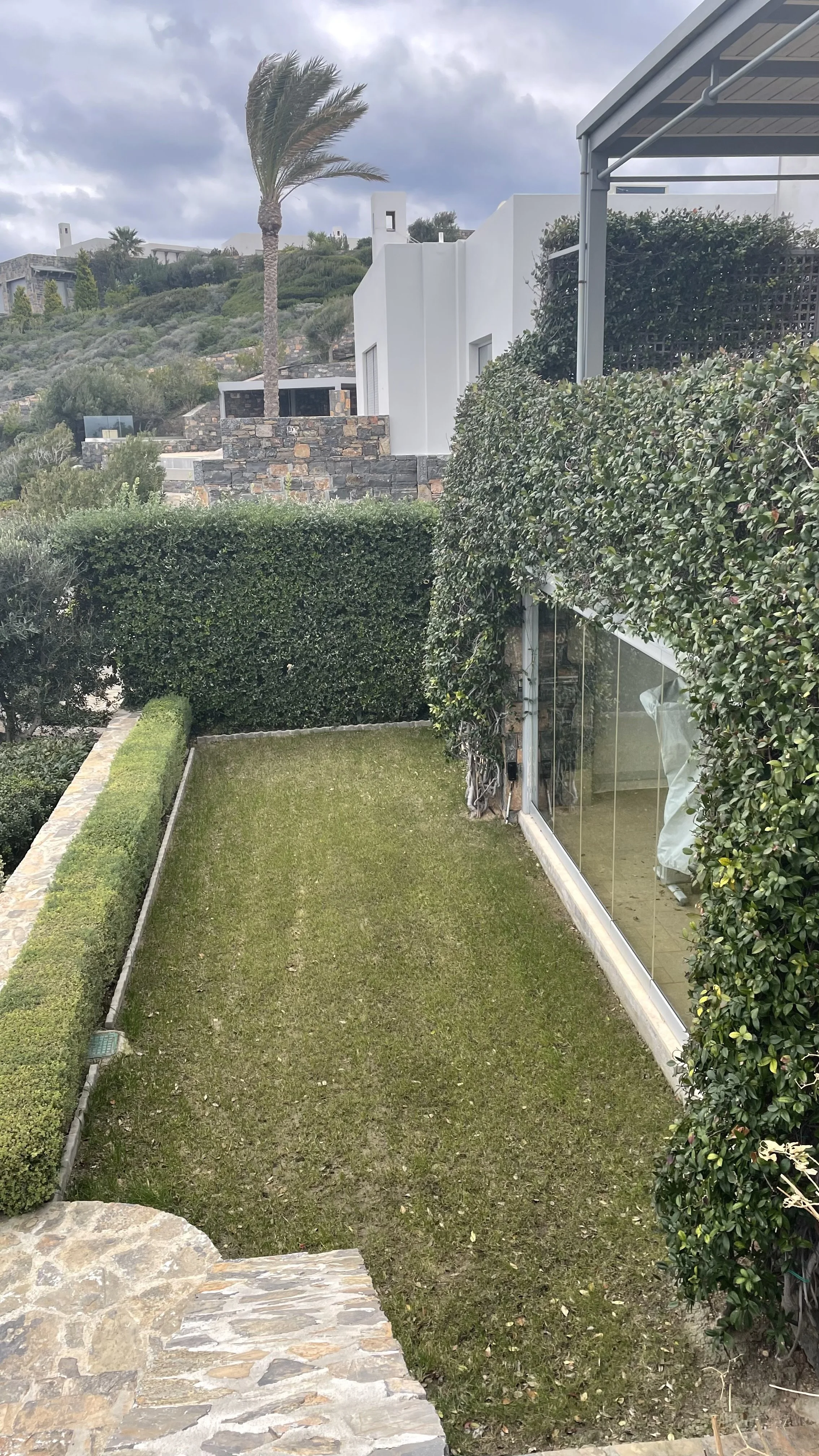 A small, well-maintained grassy backyard area with a stone border, surrounded by neatly trimmed bushes and a hedge. To the right, there is a house with large glass windows and a glass door, partially covered by foliage. In the background, a hillside 