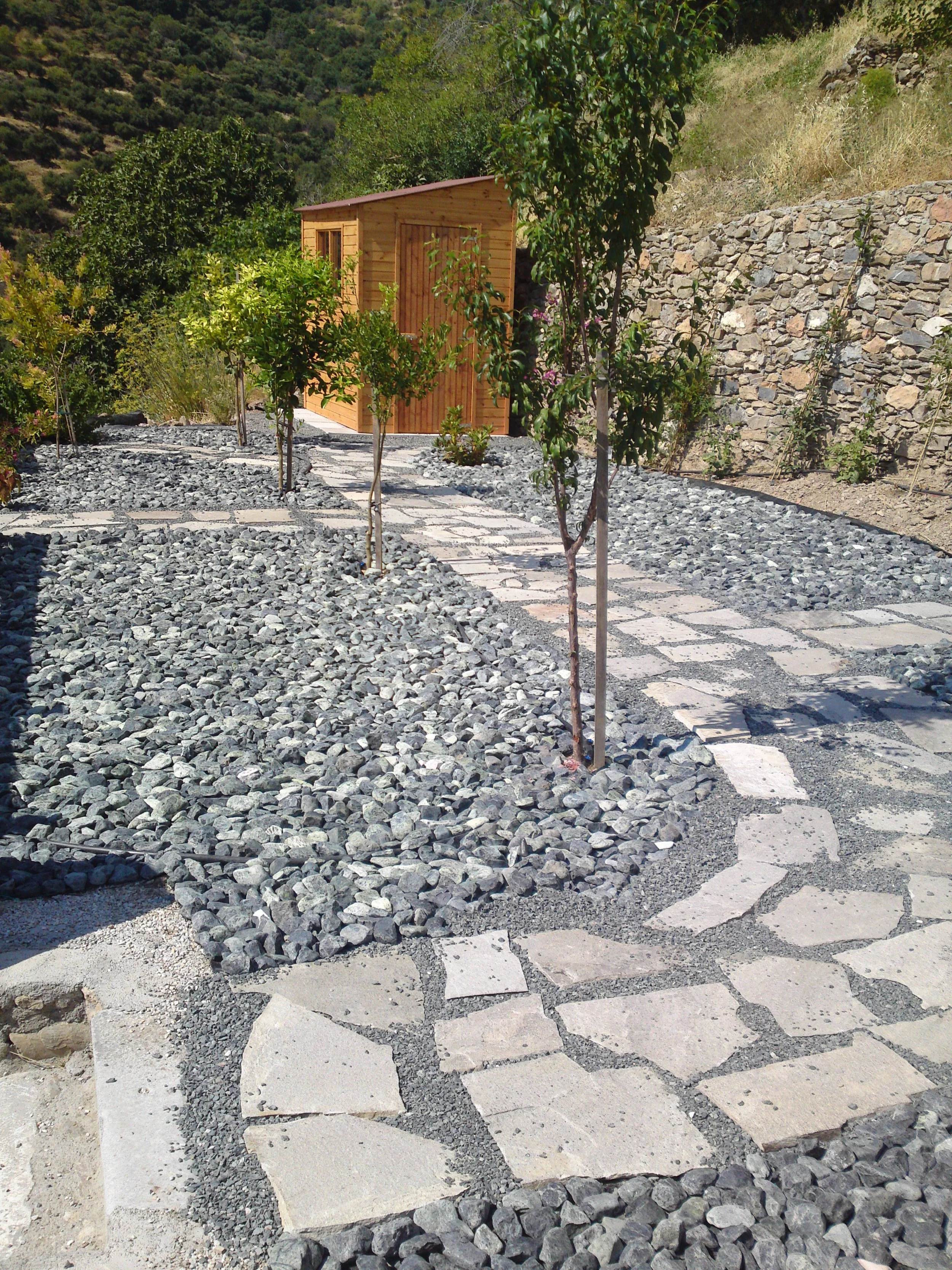 A landscaped yard with a stone and gravel pathway, young trees, and a small wooden shed on a hillside with rocky terrain and hills in the background.