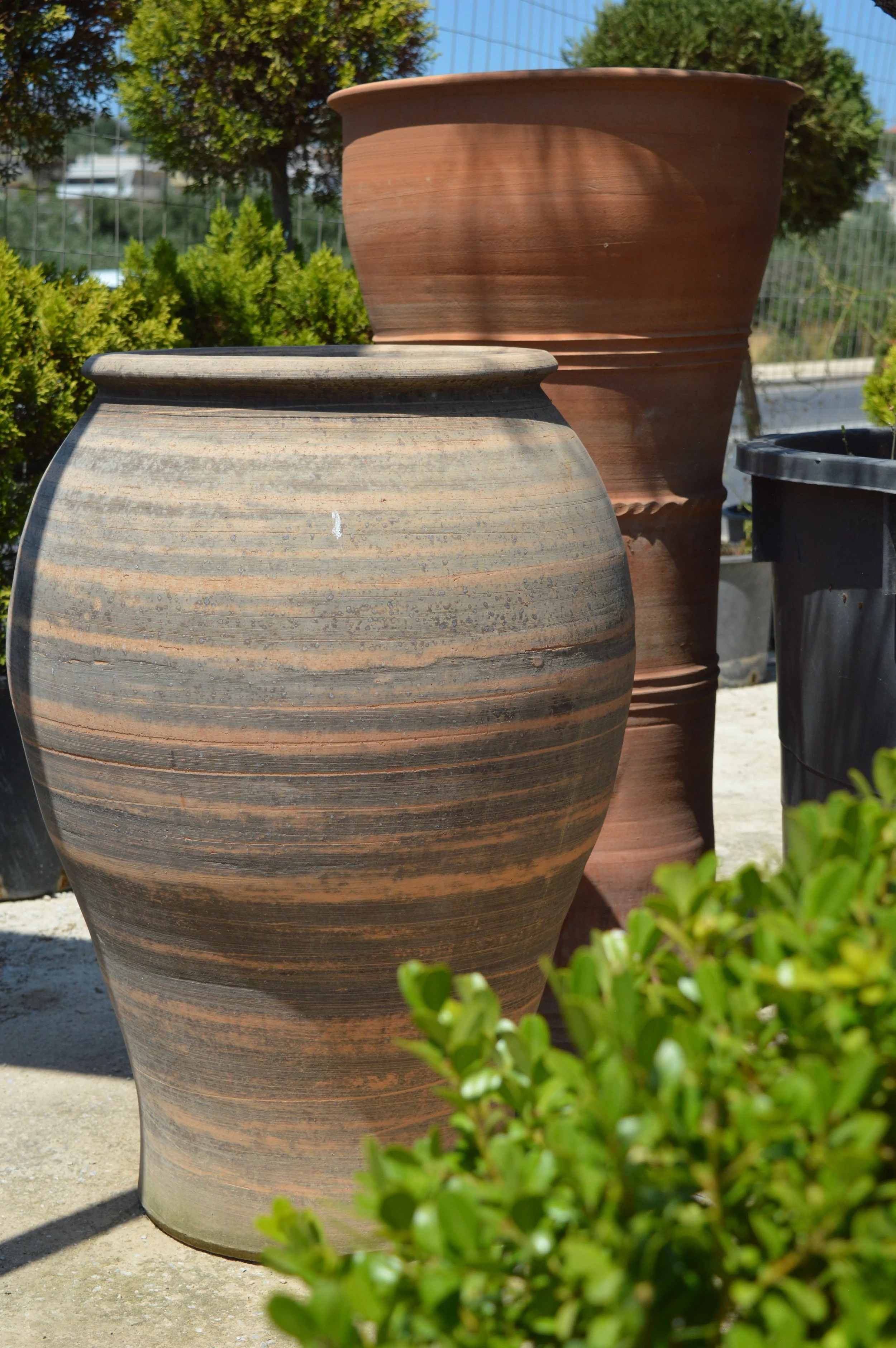 Close-up of two large terracotta garden planters outdoors, with one smaller black pot nearby, green bushes and trees in the background.