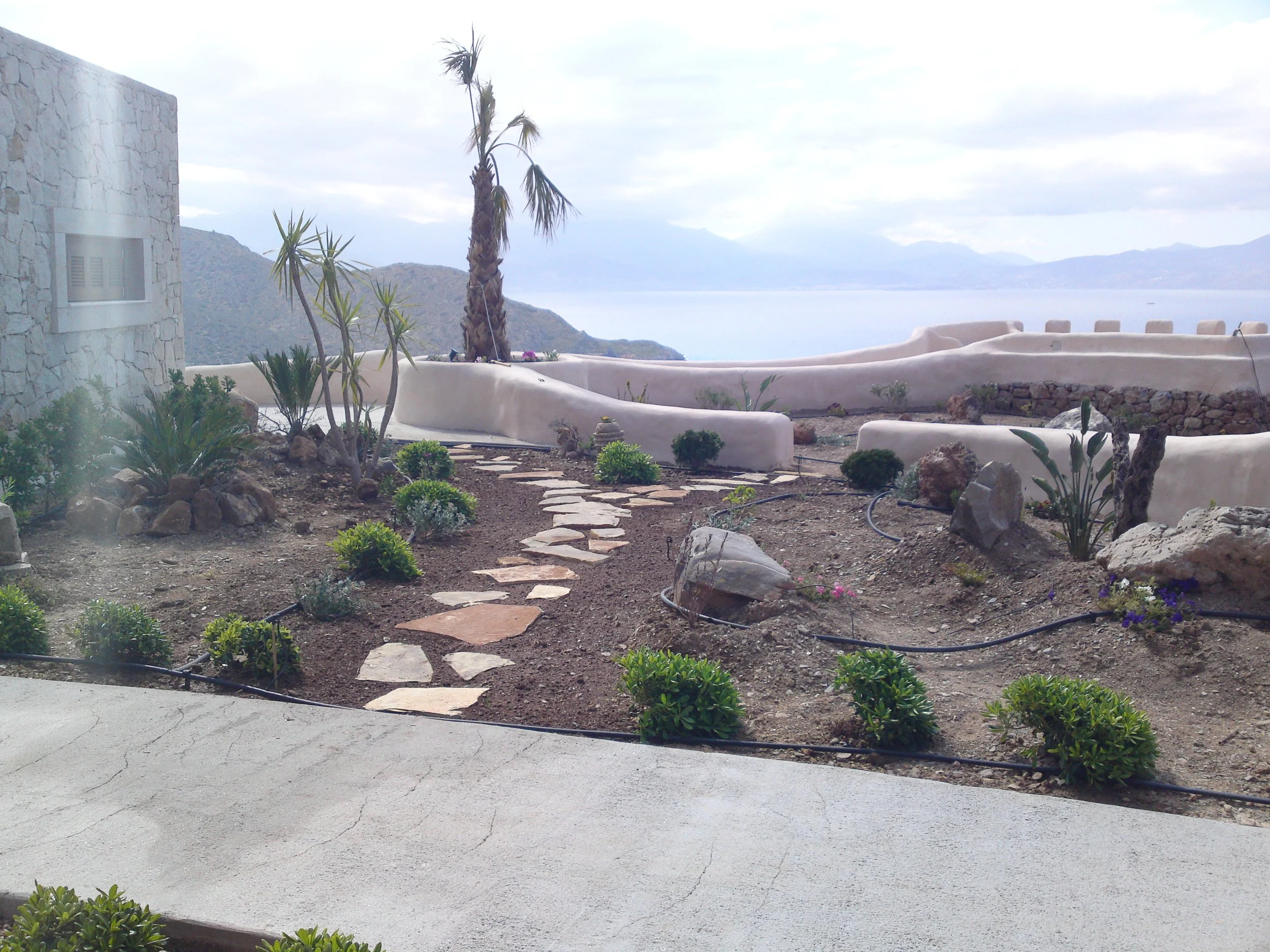 A landscaped outdoor area with desert plants and a stone pathway, overlooking a body of water and distant mountains under a cloudy sky.