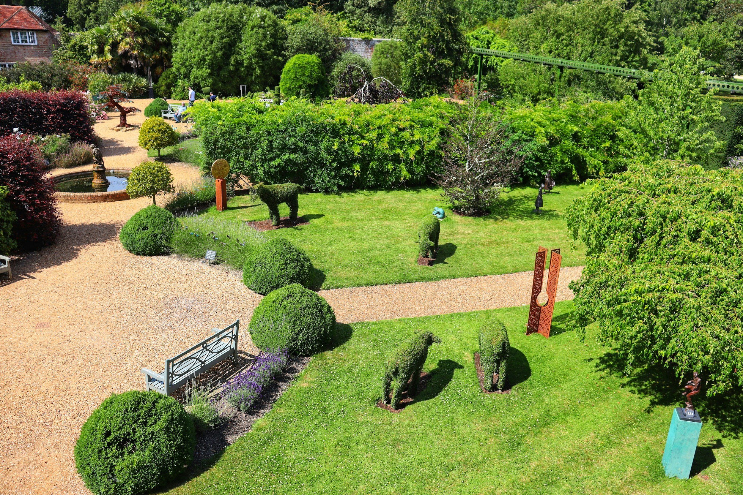 A lush, well-maintained garden with neatly trimmed bushes and trees, a small pond with a fountain, benches, and various sculptures, including a topiary dog and an abstract red sculpture, surrounded by gravel pathways and a backdrop of dense green foliage.