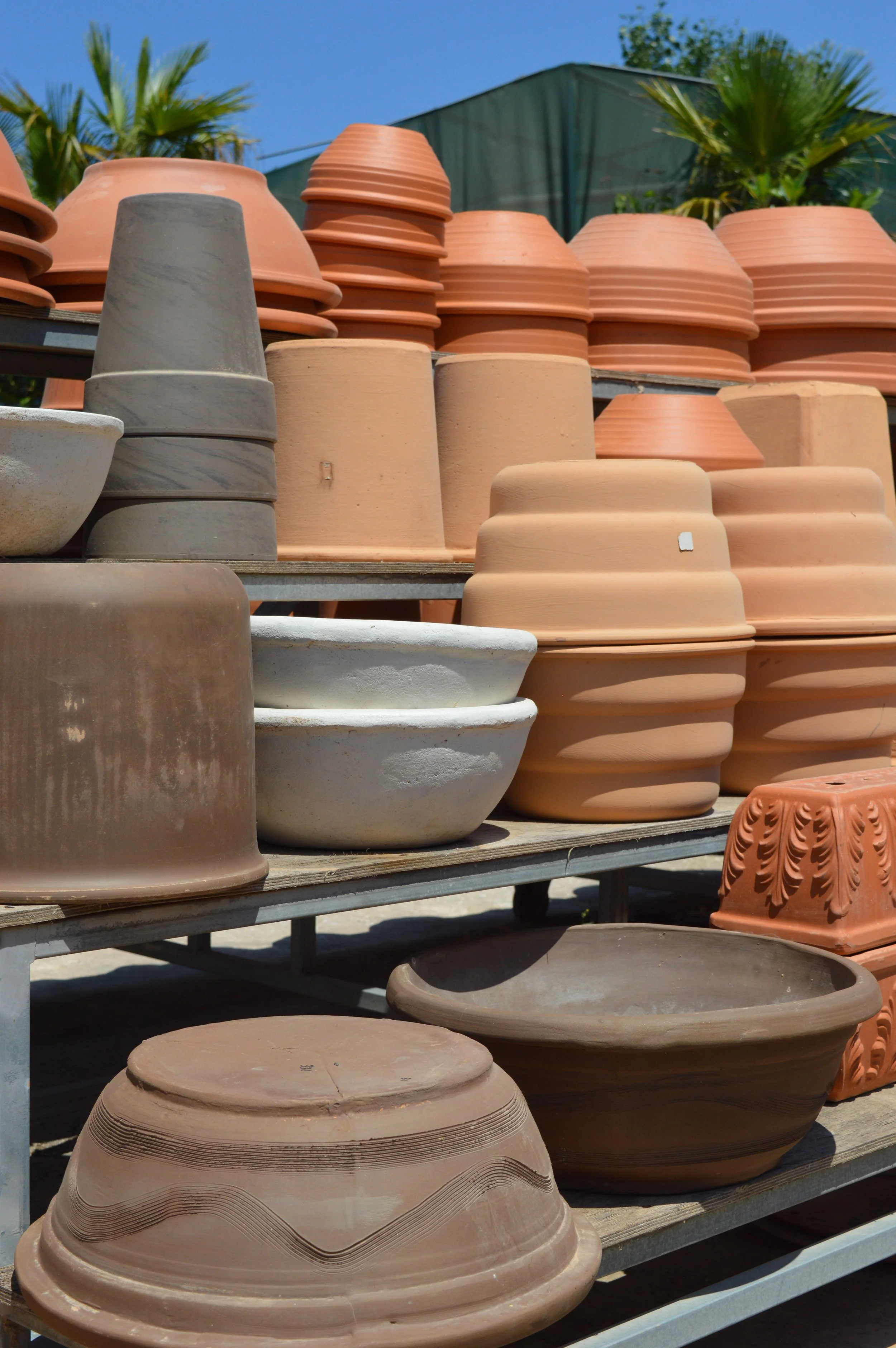 Stacks of terracotta and ceramic flower pots on metal shelves outdoors, with palm trees and blue sky in background.