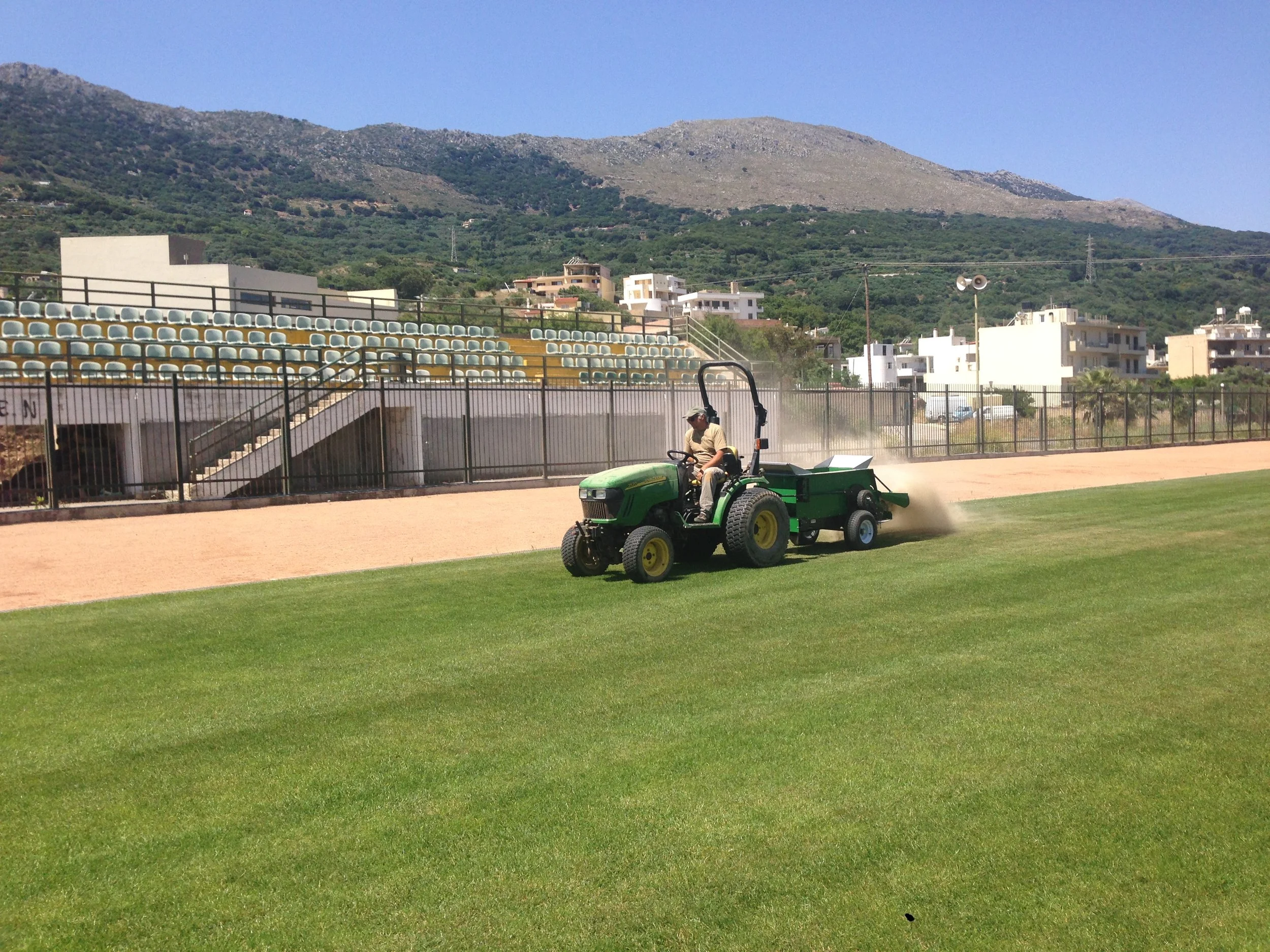 A person on a green riding lawn mower trimming grass in a sports field with empty bleacher seating, metal fencing, and a mountainous landscape in the background.