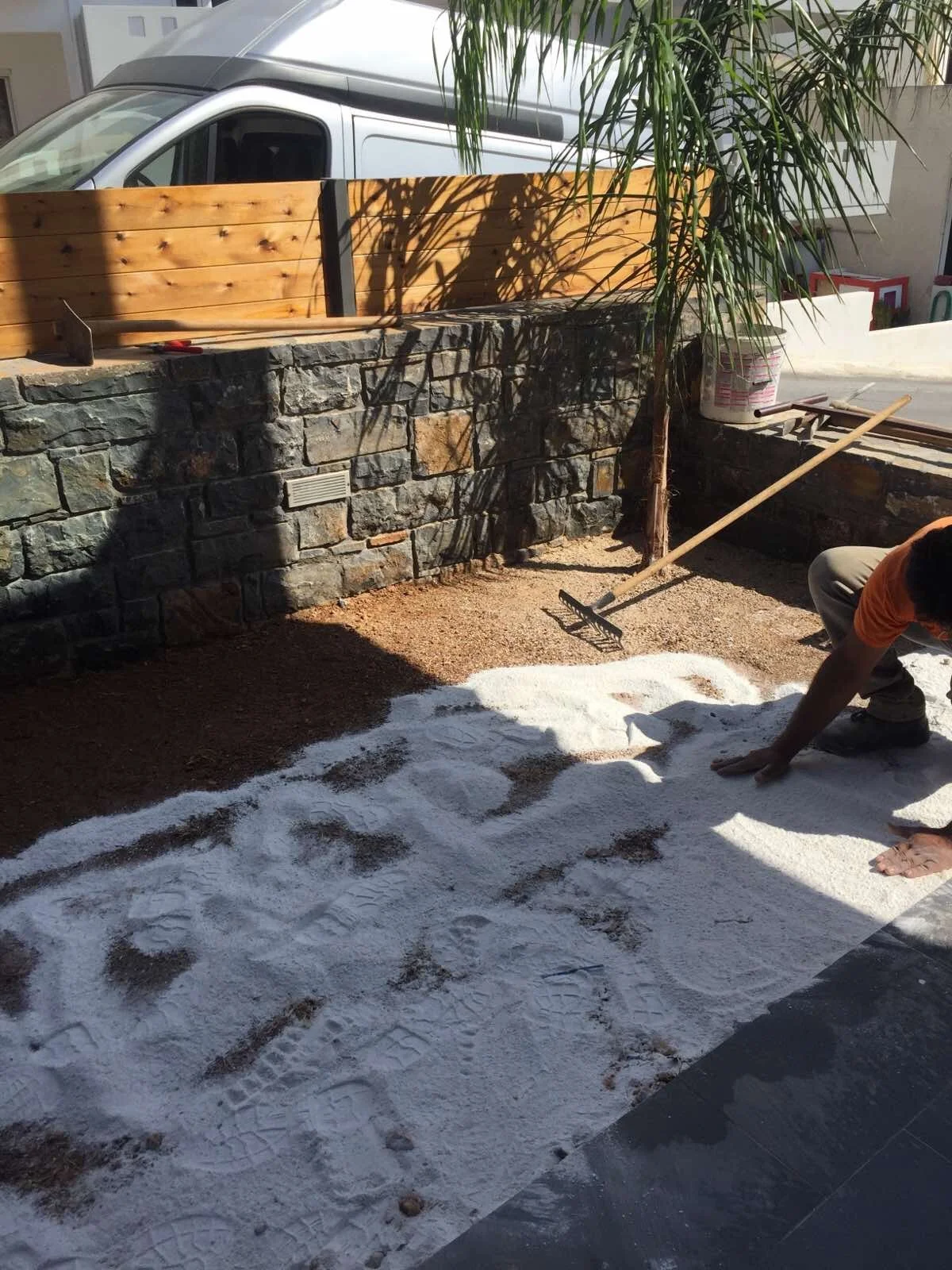 Person laying down white gravel or sand on a yard or garden area, using their hands and a rake, with a stone wall, wooden fence, and tree in the background, and a parked van nearby.