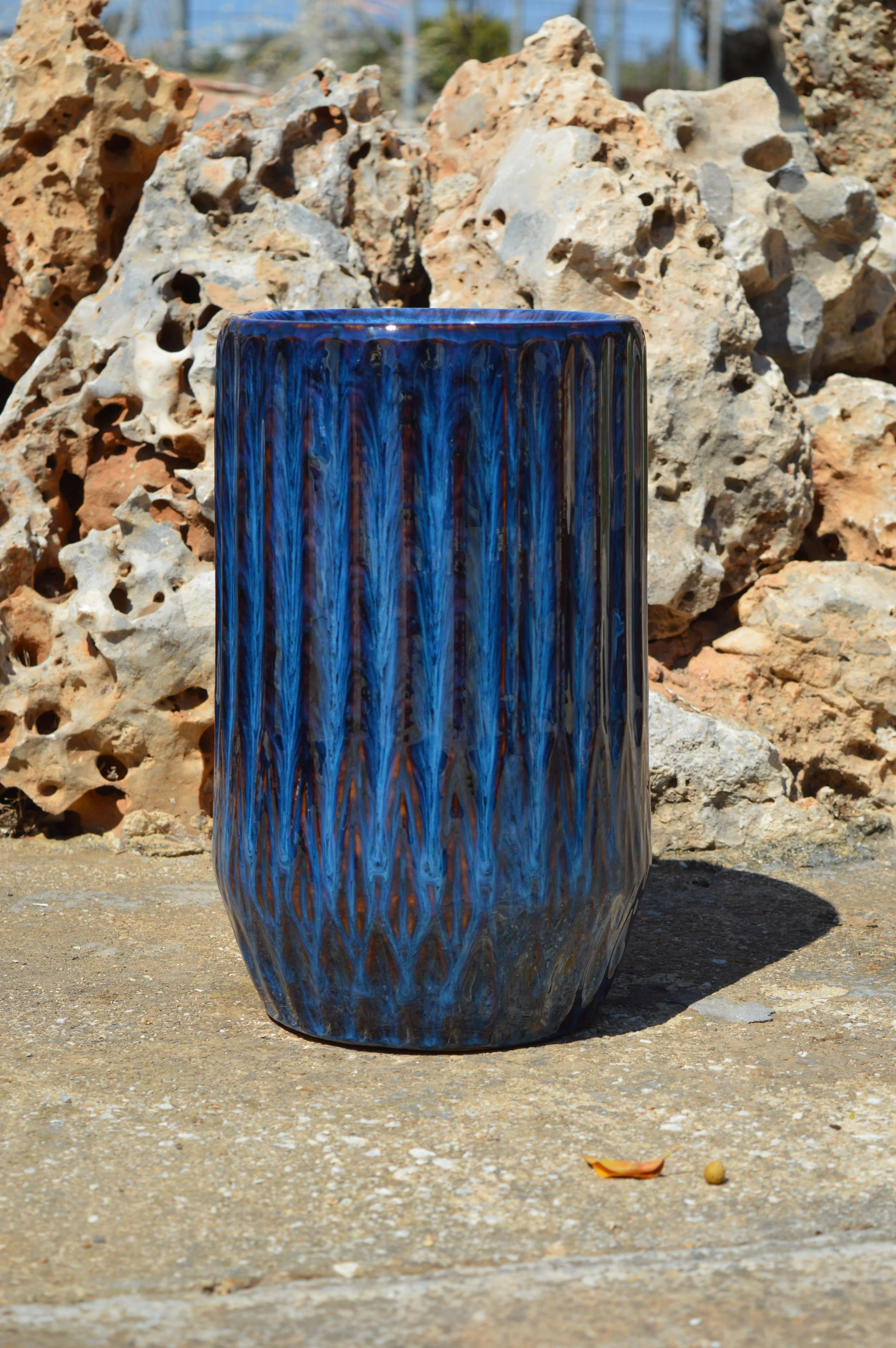 A tall, blue ceramic vase with a textured, streaked pattern is placed on a stone surface outdoors. Behind it, there is a large pile of weathered rocks with holes and crevices. The sky is clear, and trees are visible in the background.