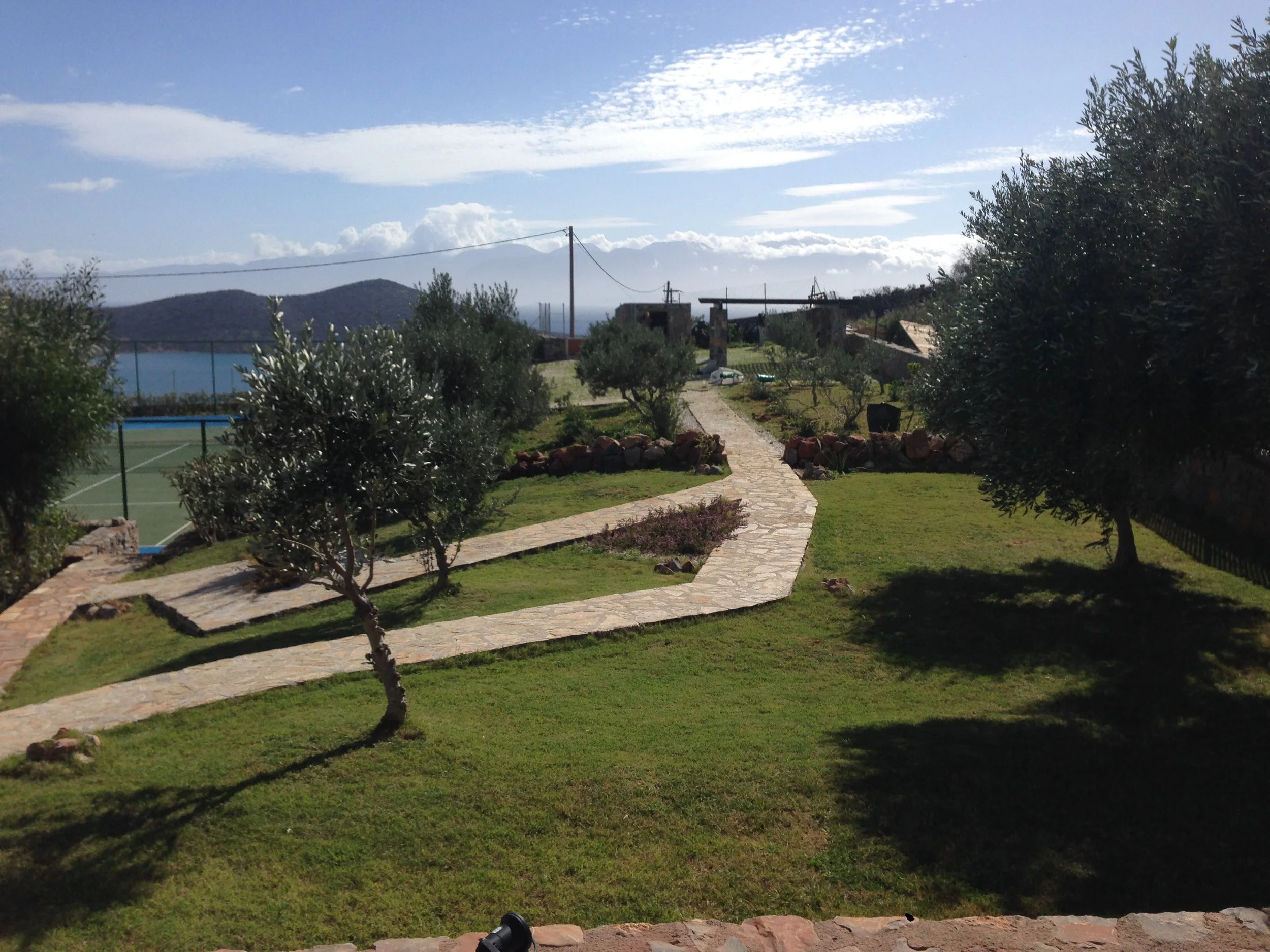 A landscaped backyard with a stone pathway, trees, and a tennis court in the distance, overlooking hills and water in the background under a partly cloudy sky.