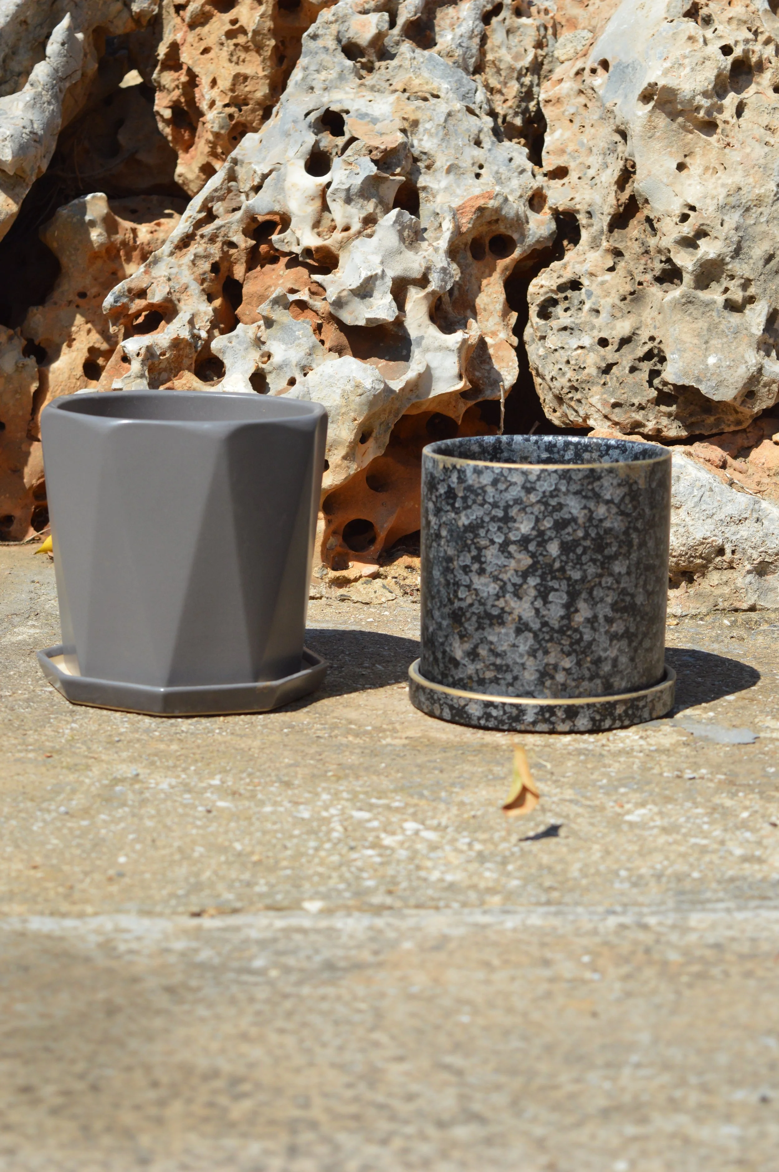 Two ceramic cups placed on a concrete surface with a rocky, porous background. The cup on the left is plain gray and resting on a saucer, while the cup on the right has a black and white speckled pattern.