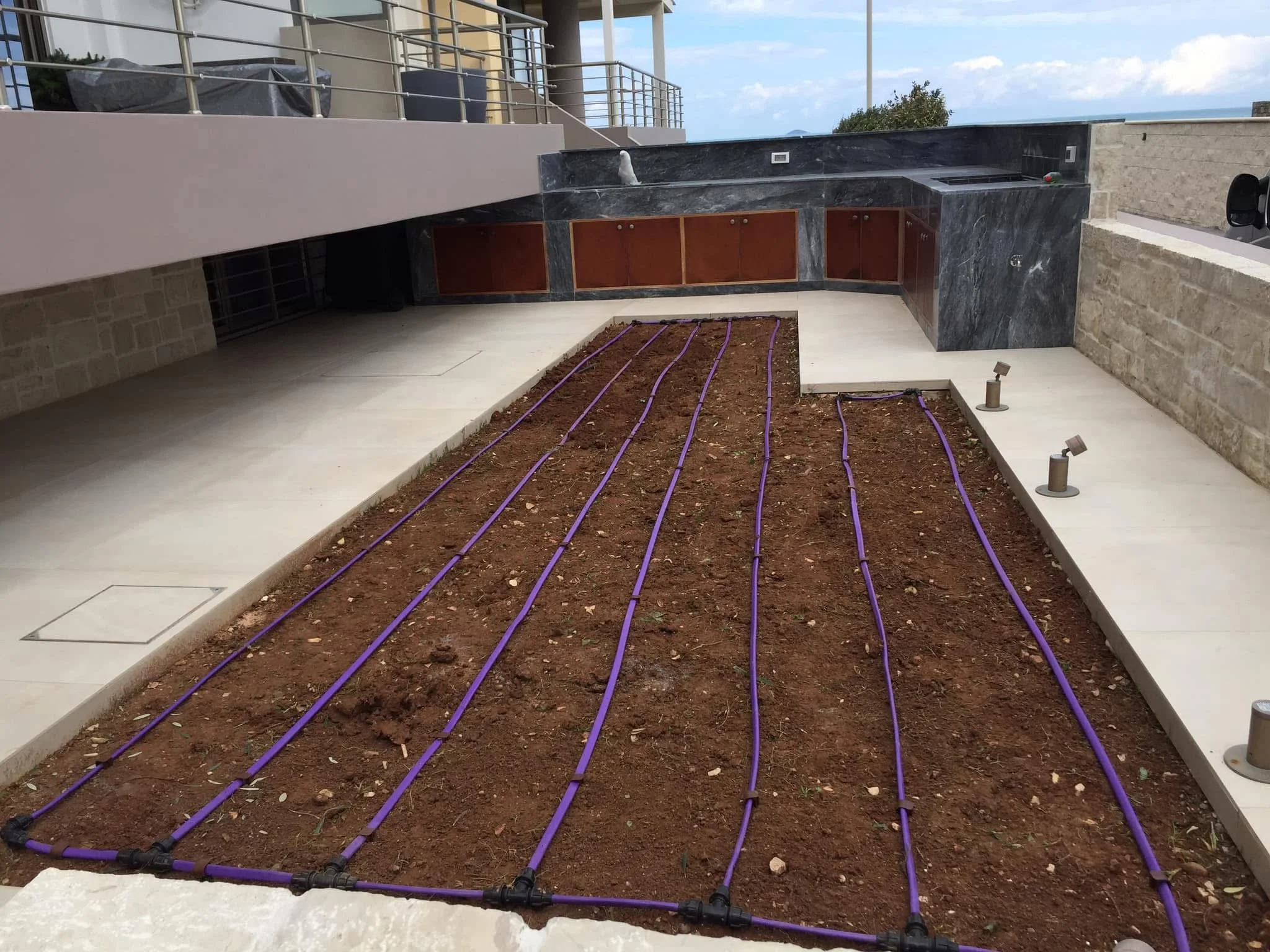 Construction site with purple irrigation pipes laid out on soil in a patio area, adjacent to kitchen cabinets and granite countertop, with a view of a balcony and the ocean in the background.