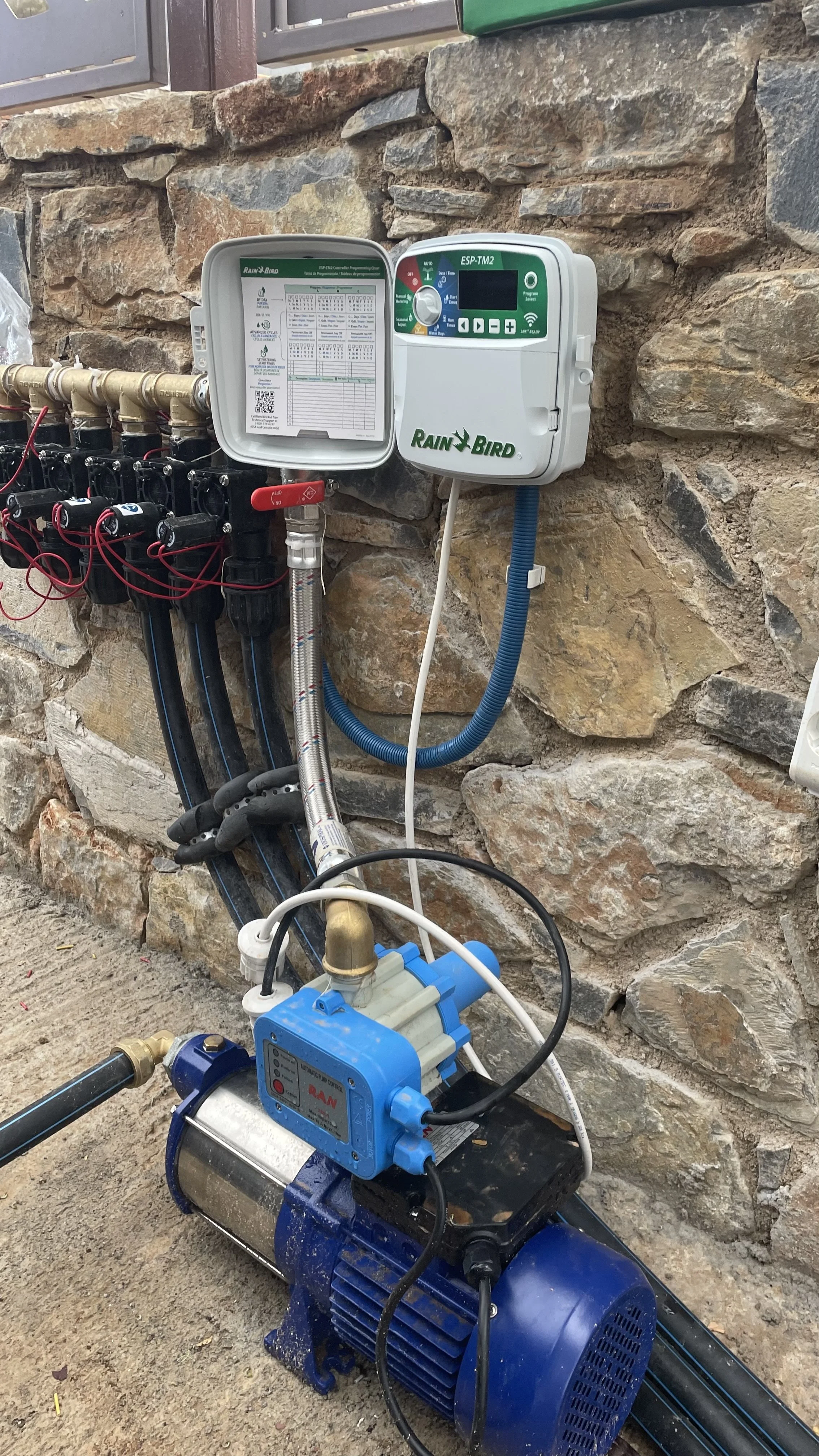An outdoor setup of irrigation equipment including a Rain Bird controller, a blue pump, and various pipes and fittings against a stone wall.