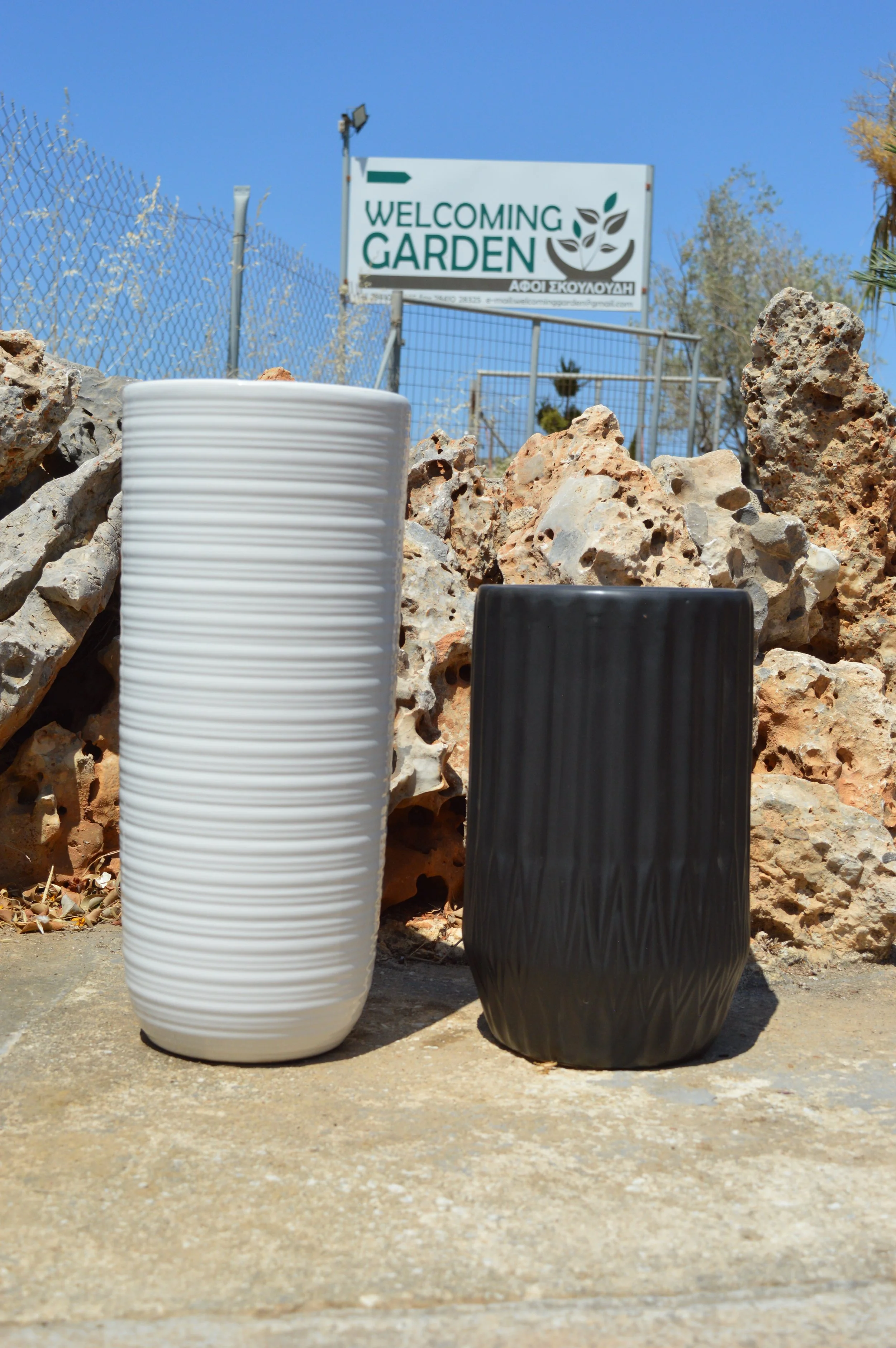 Two ceramic vases, one white and tall, the other black and shorter, placed on a concrete surface in front of a rocky background with a fencing and a sign that reads 'Welcoming Garden' with a leaf logo, under a clear blue sky.