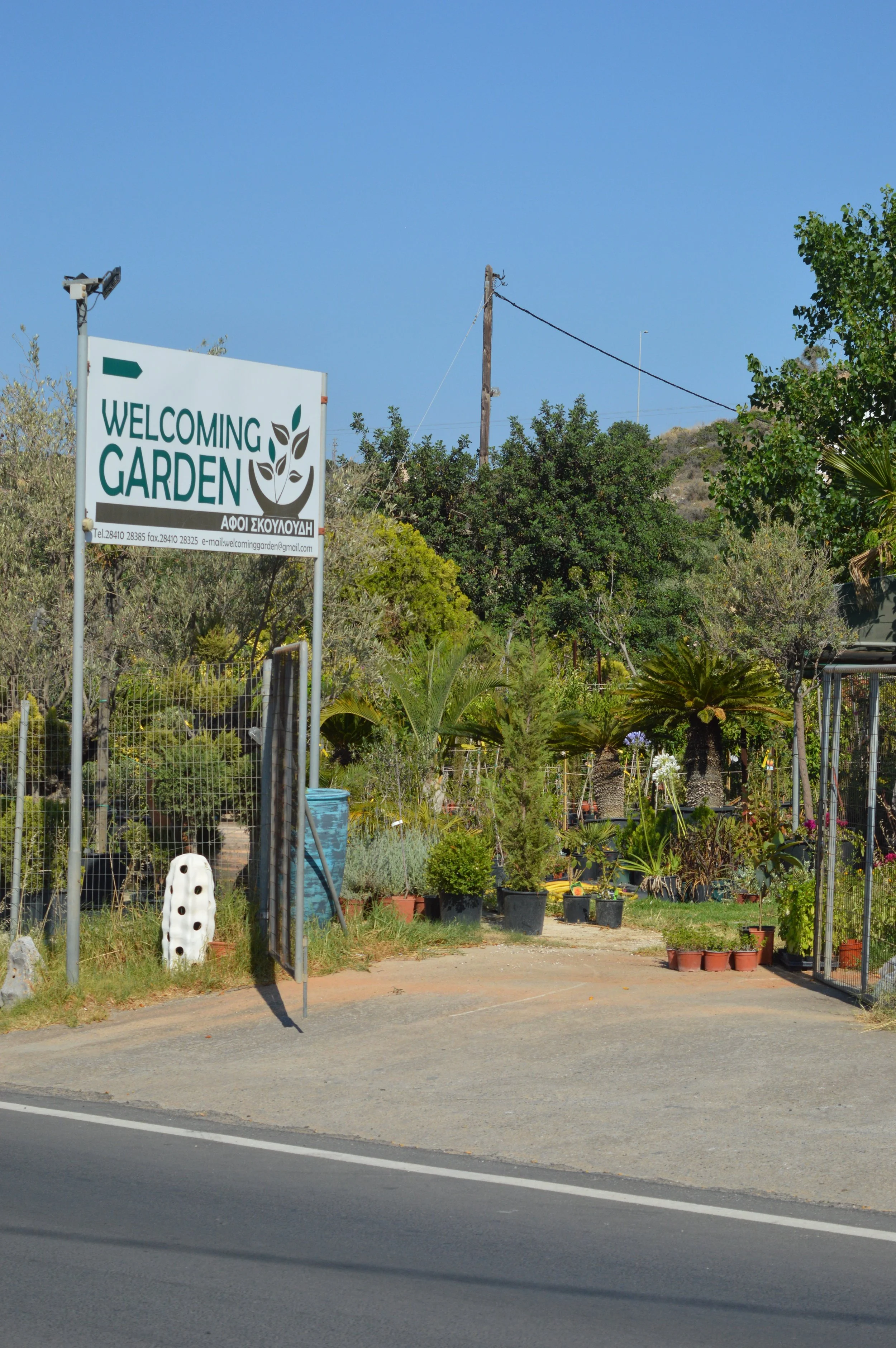 A garden center with various potted plants and trees, a sign that reads 'Welcoming Garden' with contact information, and a blue sky overhead.