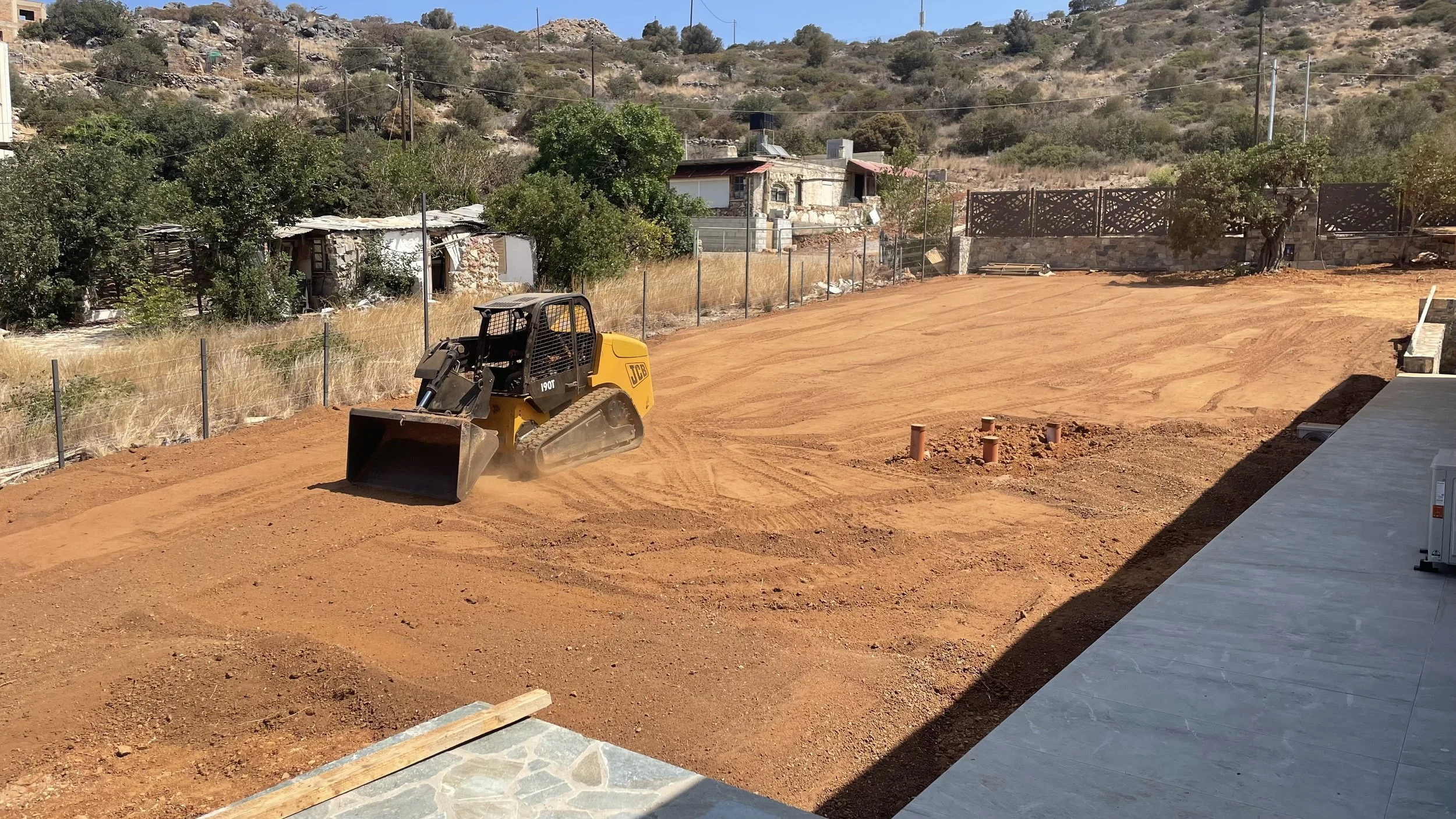 A construction site with a yellow JCB compact excavator moving reddish dirt, surrounded by a chain-link fence, with a house and trees in the background and a hill with sparse vegetation under a clear blue sky.
