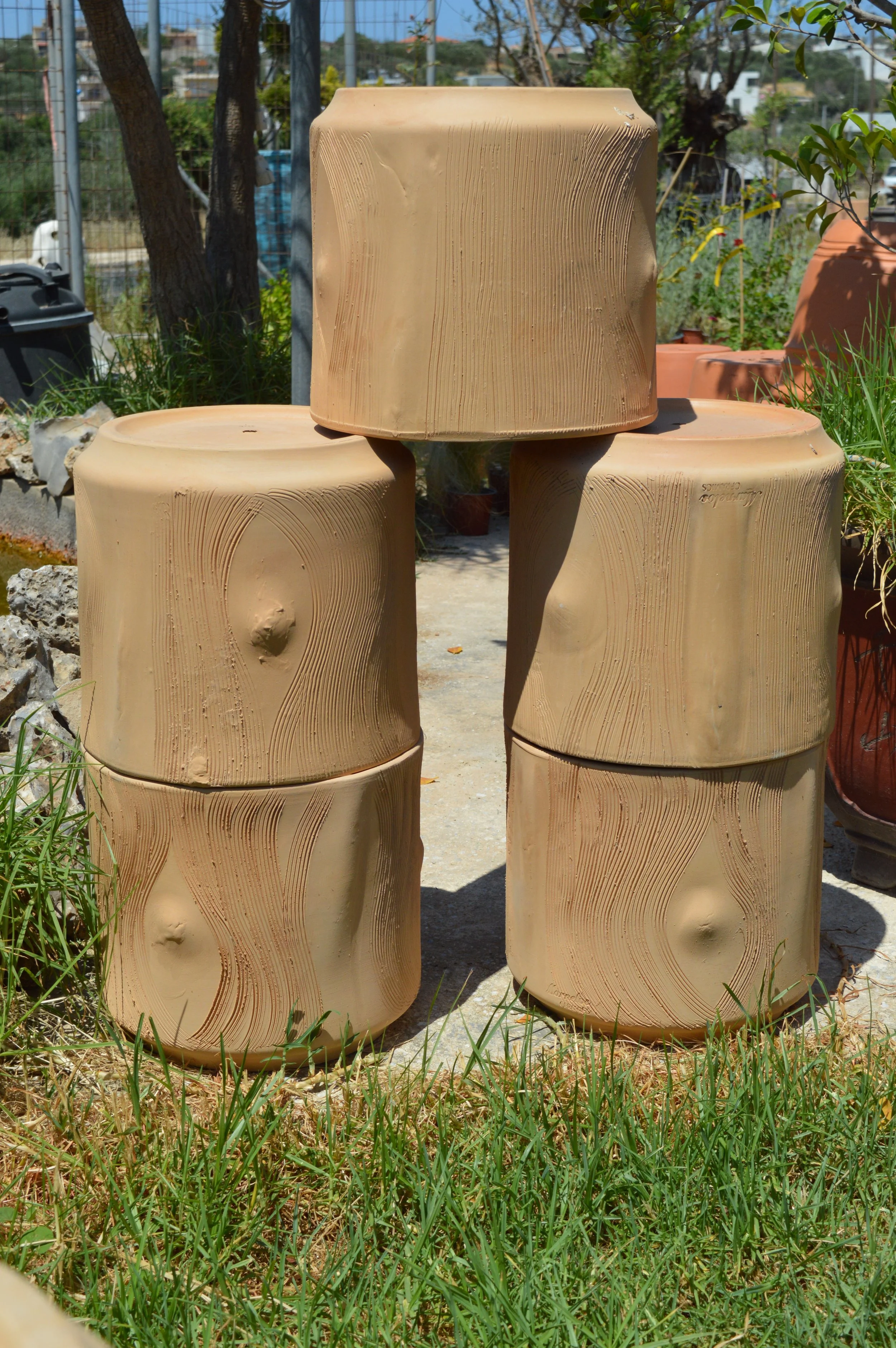 Multiple beige clay pots with textured wood grain patterns stacked outdoors on grass, with trees and a fence in the background.