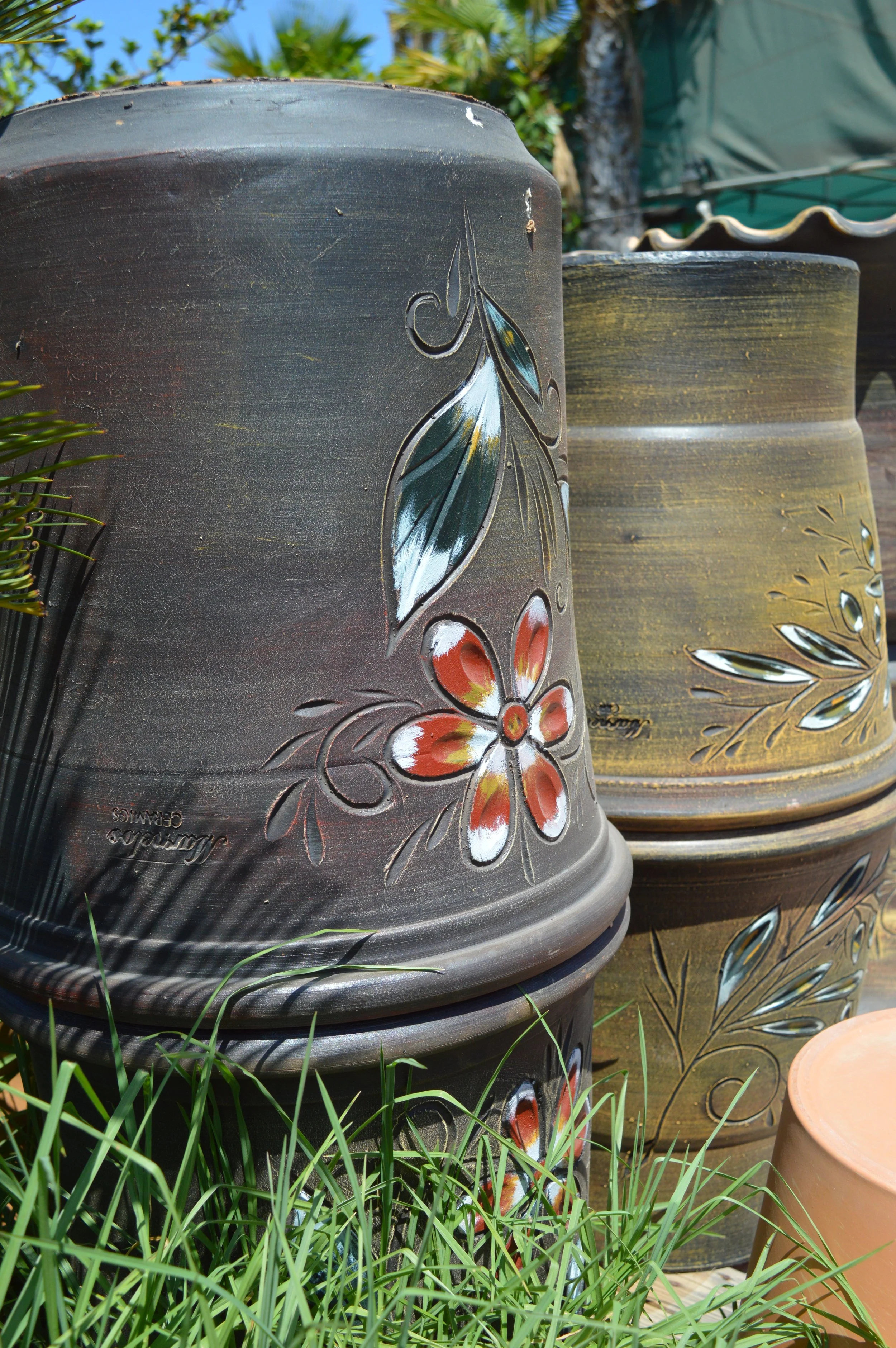 Close-up of black ceramic flower pots with colorful floral and leaf designs, placed outdoors among grass and greenery.